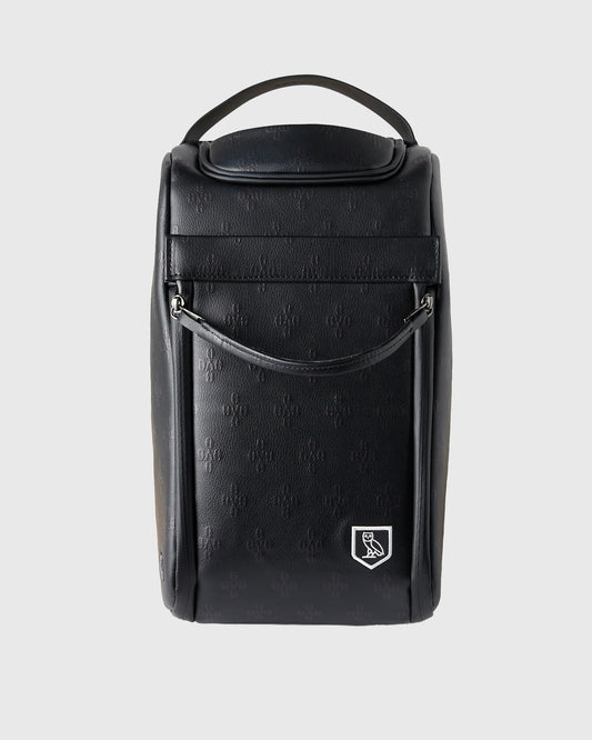 Black cylindrical golf shoe bag with handle, monogram texture, and crest logo on front.