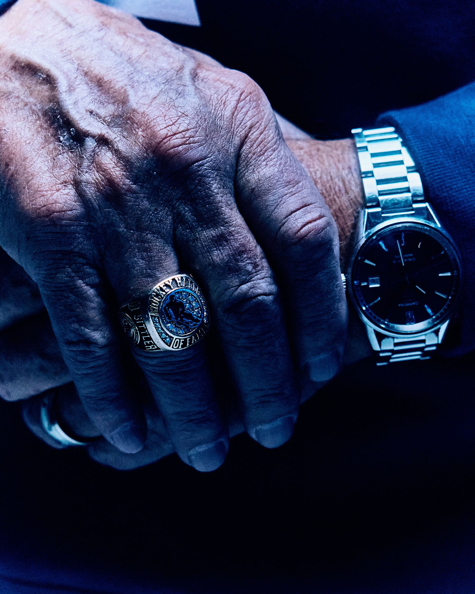 Close-up of a Darryl Sittler's hands wearing a silver ring and a watch, with a dark background.