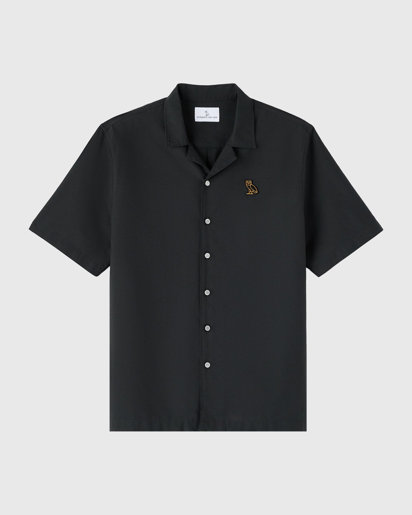 Black short-sleeve camp shirt with white buttons and small embroidered owl logo on chest.