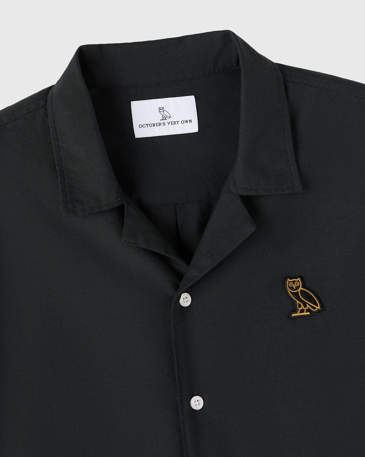 Black camp shirt with notched collar, white buttons, and gold owl embroidery on chest.