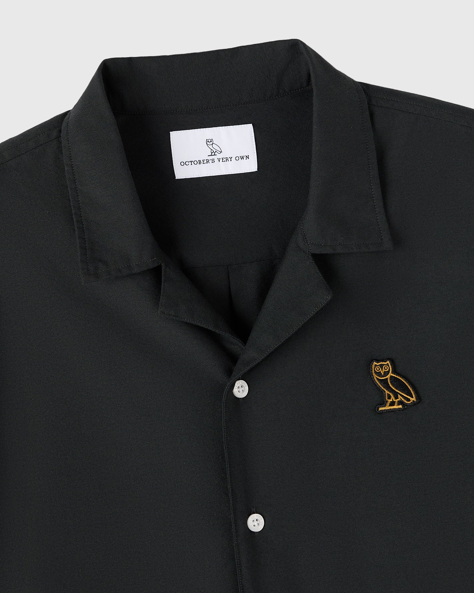 Black camp shirt with notched collar, white buttons, and gold owl embroidery on chest.