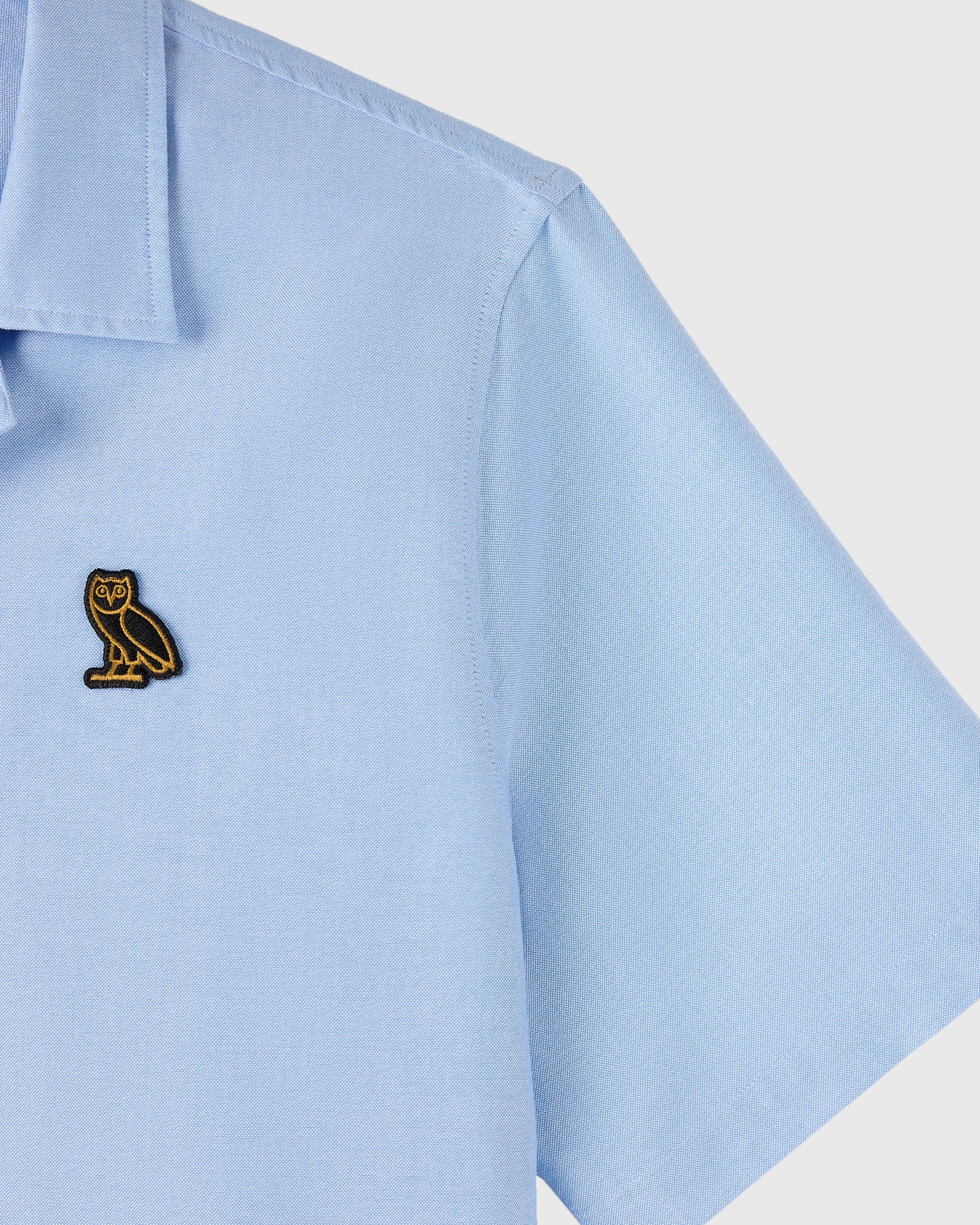 Light blue oxford camp shirt with short sleeves, chest pocket, and small gold owl embroidered logo.