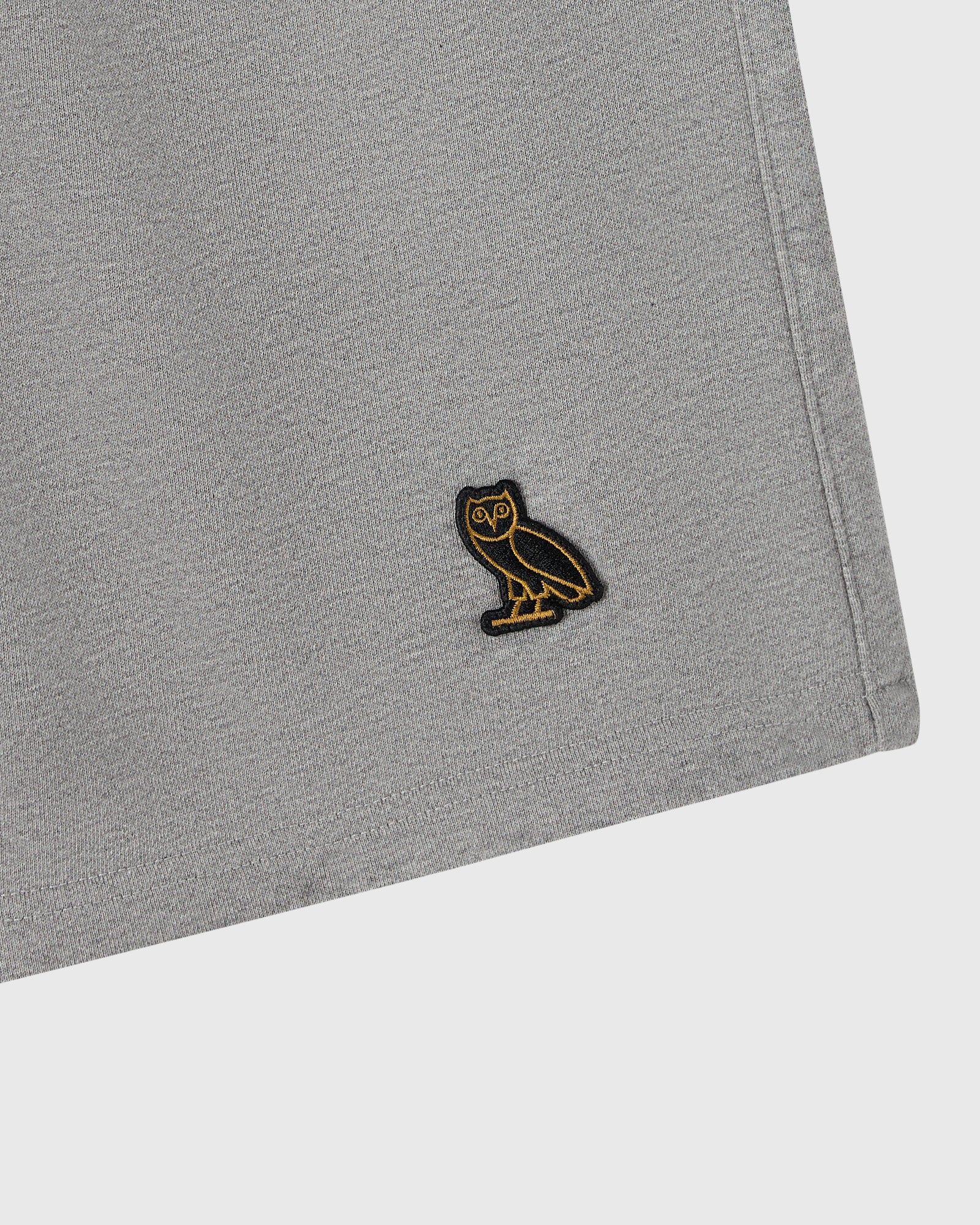 Heather grey sweatshort with small black and gold owl embroidered patch on thigh.