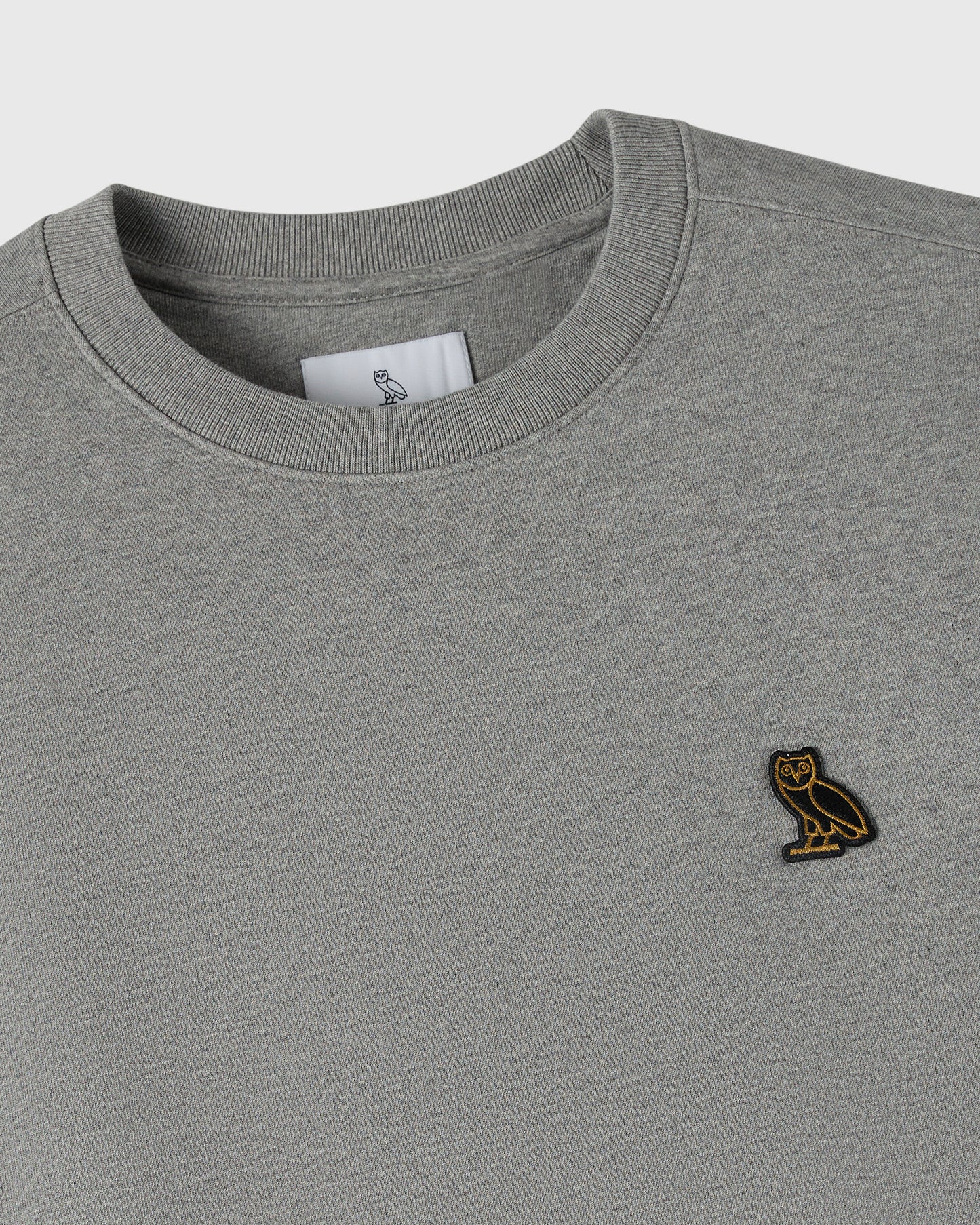 Heather grey crewneck sweatshirt with ribbed neckline and small owl embroidered patch on chest.
