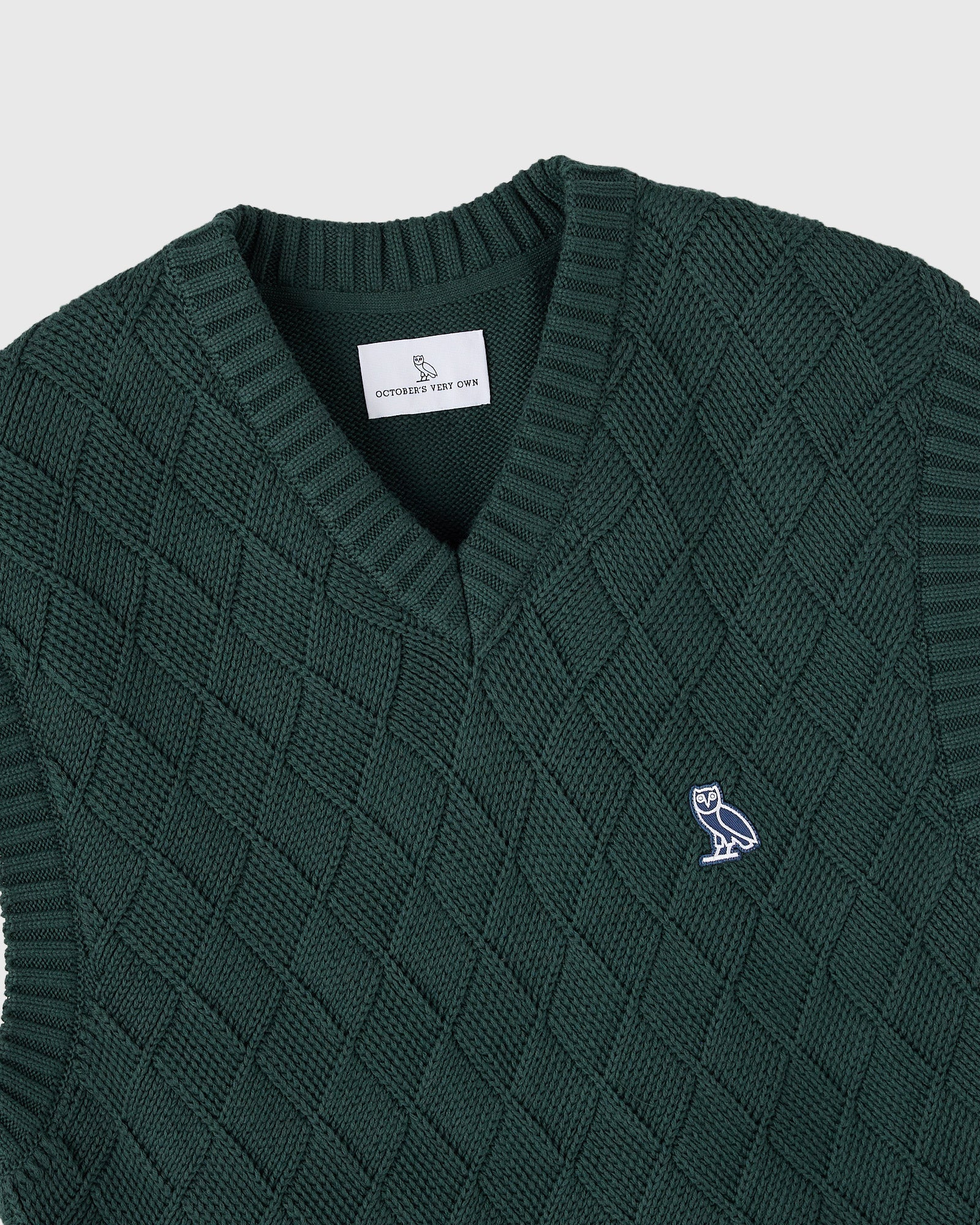 Midnight green basketweave sweater vest with V-neckline, ribbed collar and armholes, and embroidered owl logo.