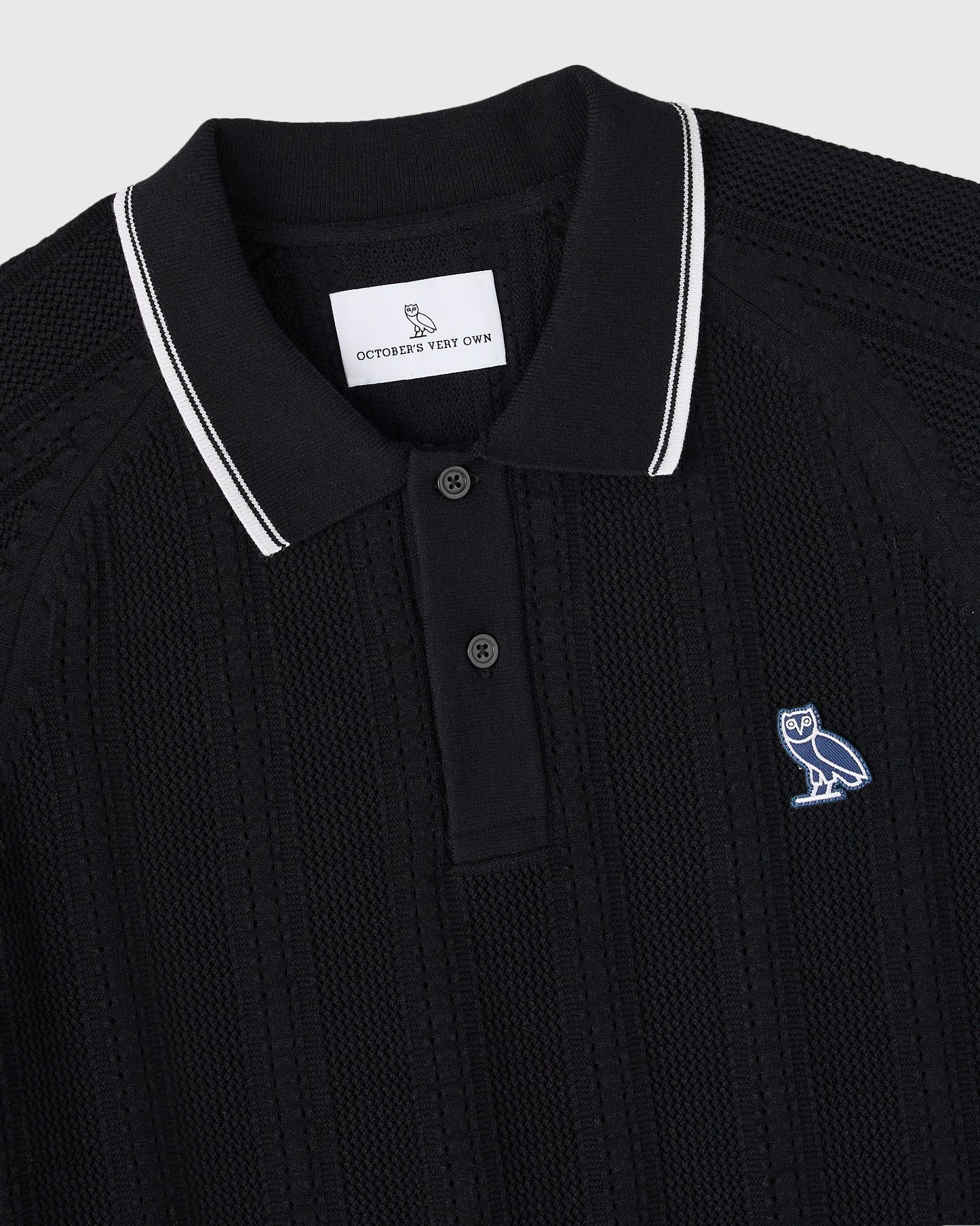 Black open knit polo sweater with white striped collar and small embroidered owl logo on chest.