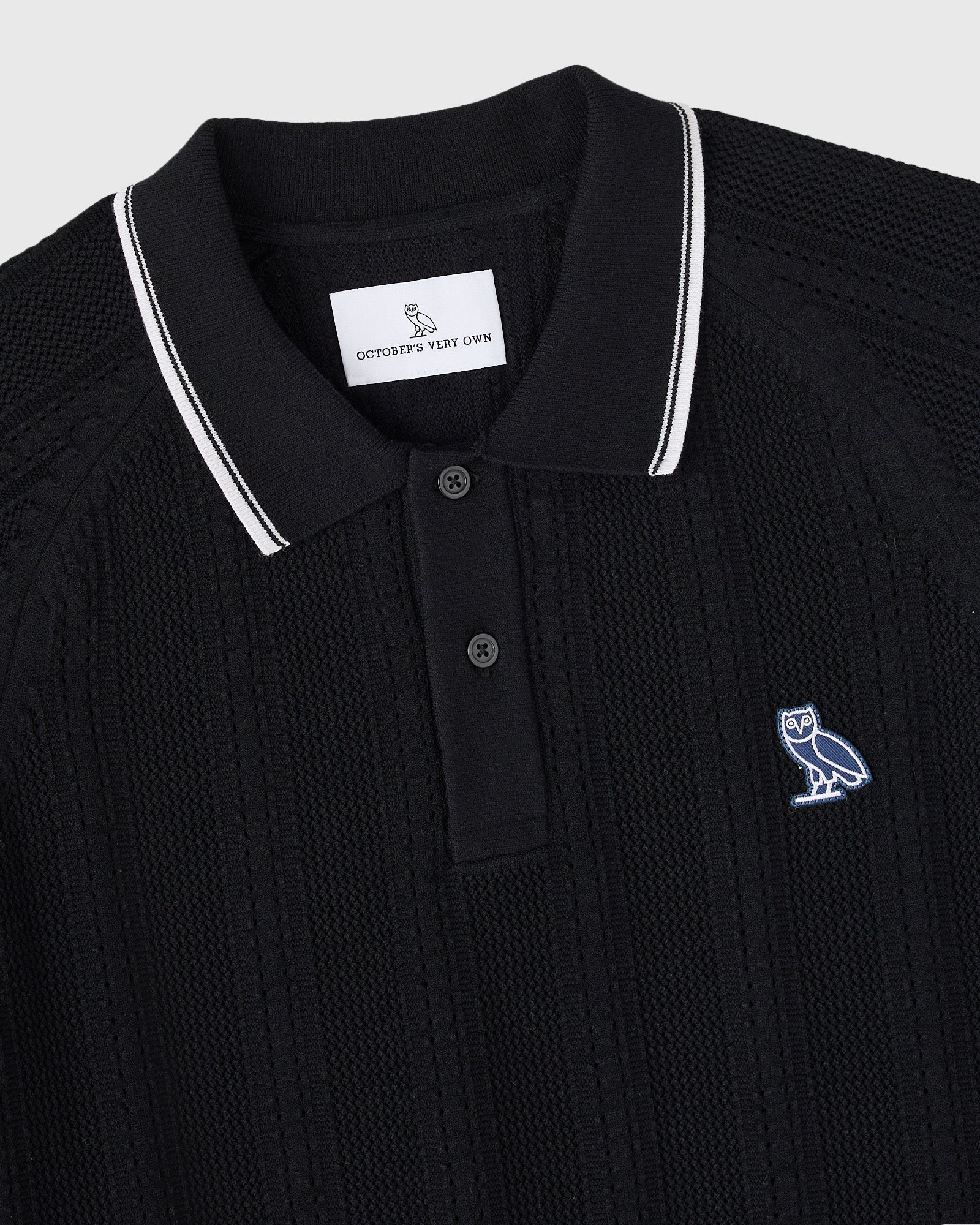 Black open knit polo sweater with white striped collar and small embroidered owl logo on chest.