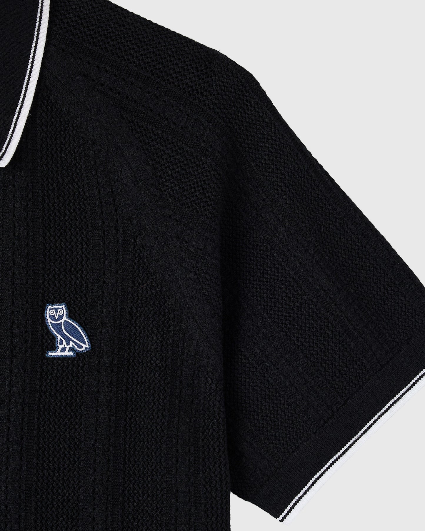 Black open knit sweater polo with white trim and embroidered owl logo on chest.
