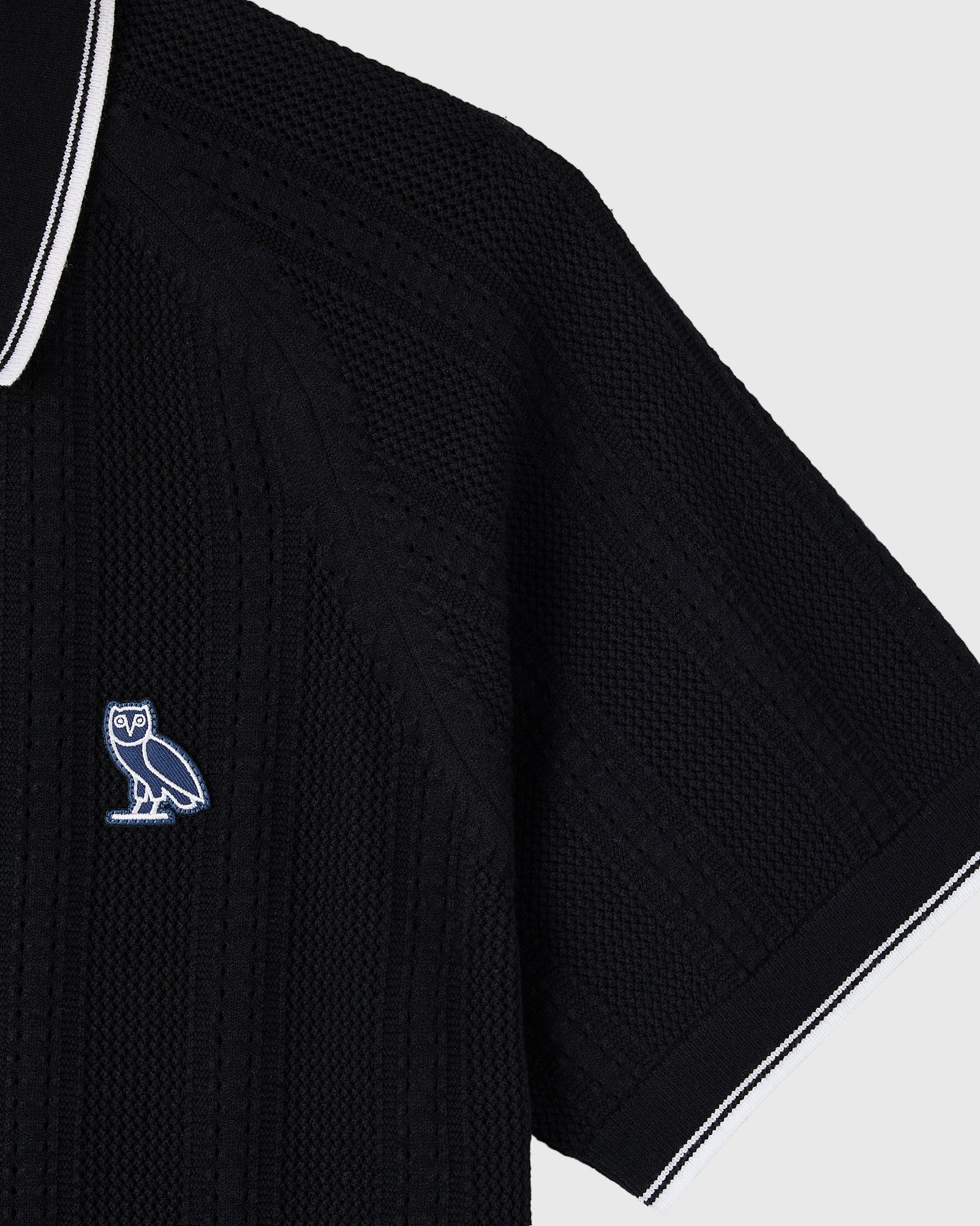 Black open knit sweater polo with white trim and embroidered owl logo on chest.