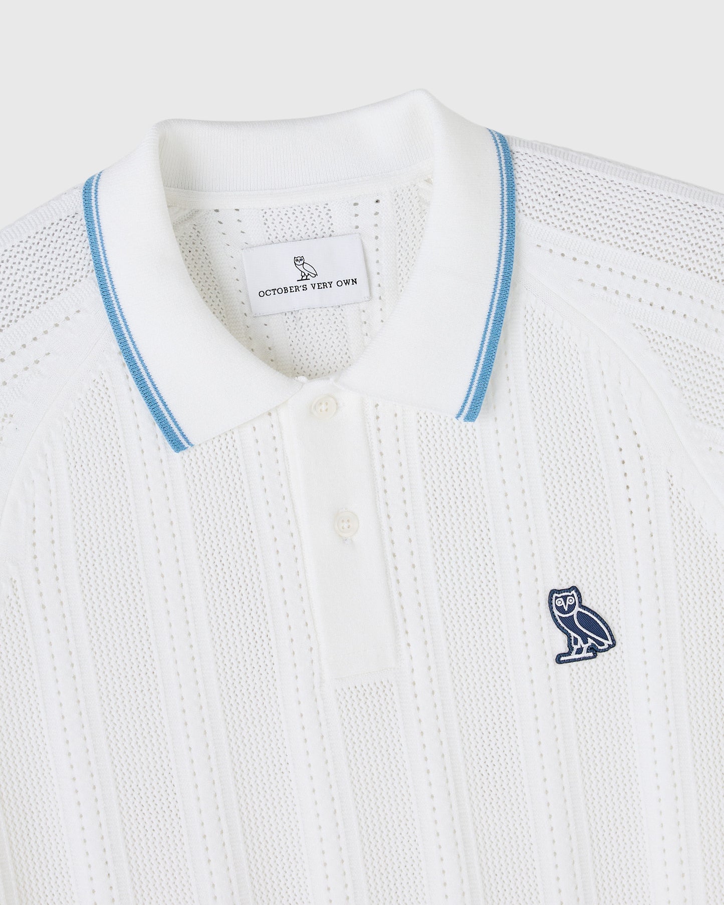 White open knit polo sweater with blue striped collar trim and navy owl emblem on chest.