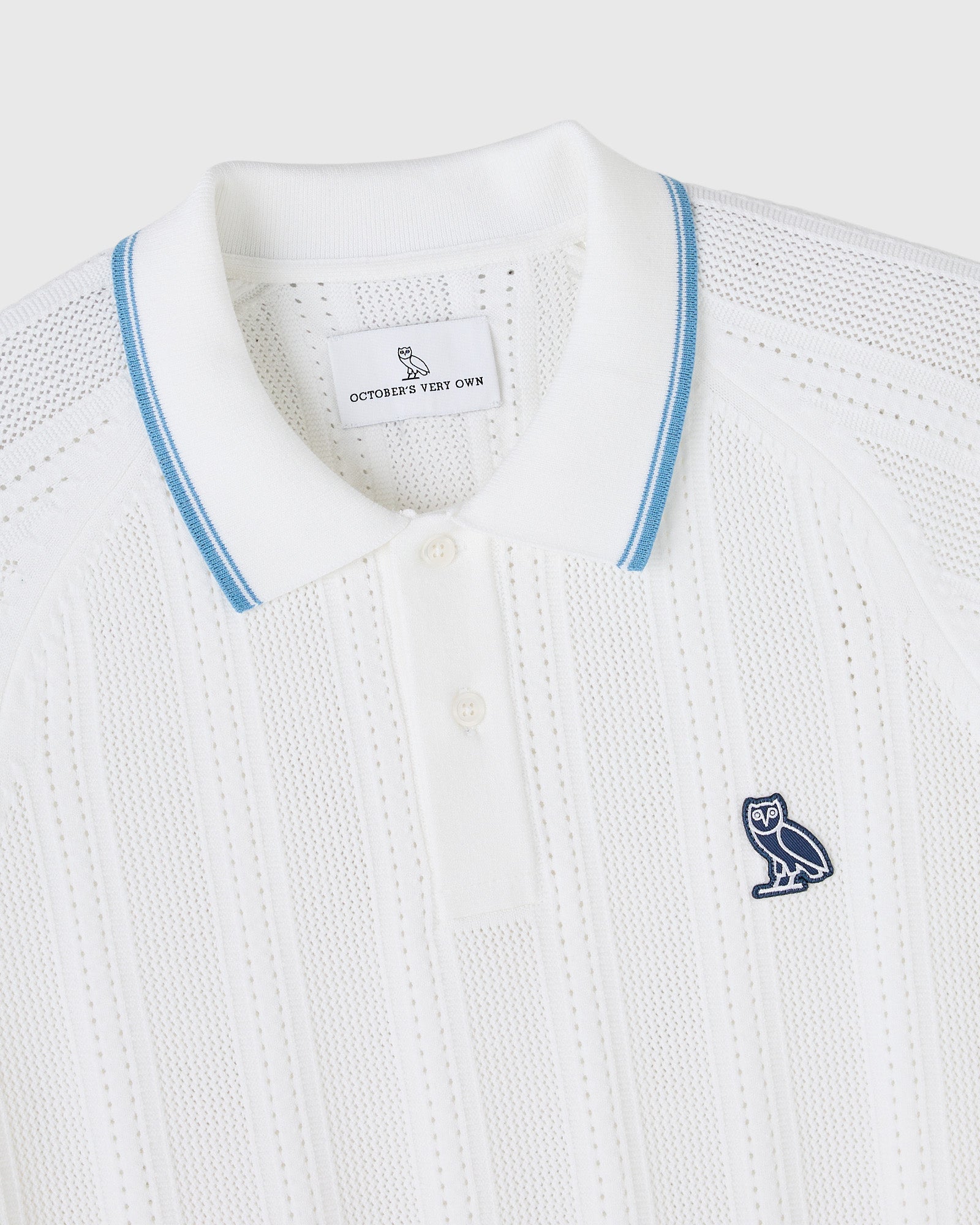 White open knit polo sweater with blue striped collar trim and navy owl emblem on chest.