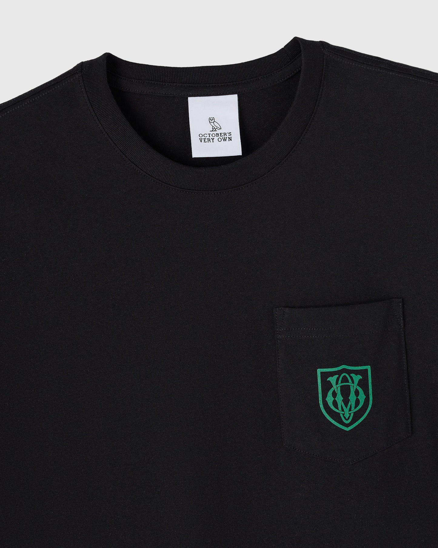 Black t-shirt with OVO owl crest logo in green on chest pocket and brand label at neckline.