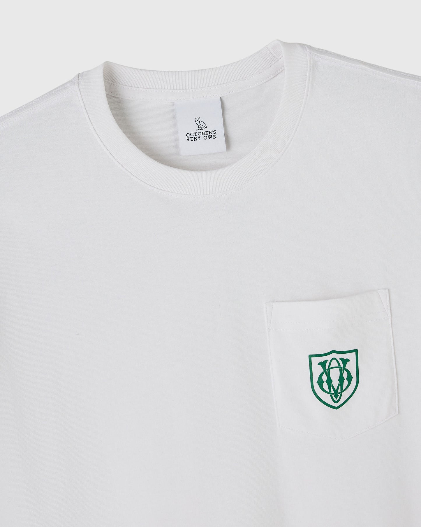 White crewneck t-shirt with OVO owl shield logo embroidered in green on chest pocket and brand tag at neck.