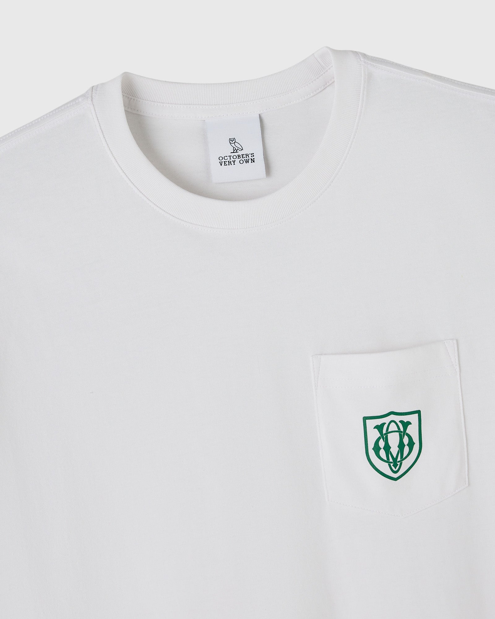 White crewneck t-shirt with OVO owl shield logo embroidered in green on chest pocket and brand tag at neck.