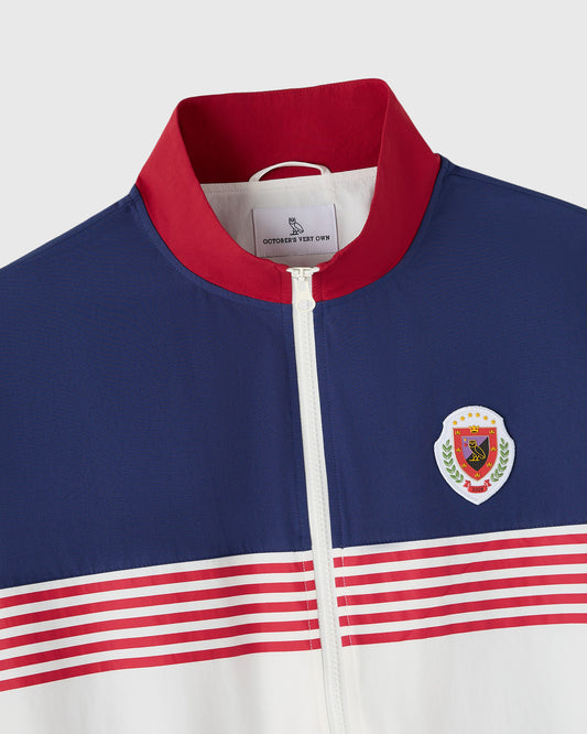 Deep denim nylon track jacket with red collar, white zip front, embroidered crest, and red-white striped lower panels.