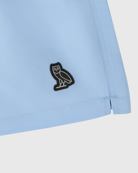 Light blue swim short with black owl embroidered patch on hip and visible drawstring detail.