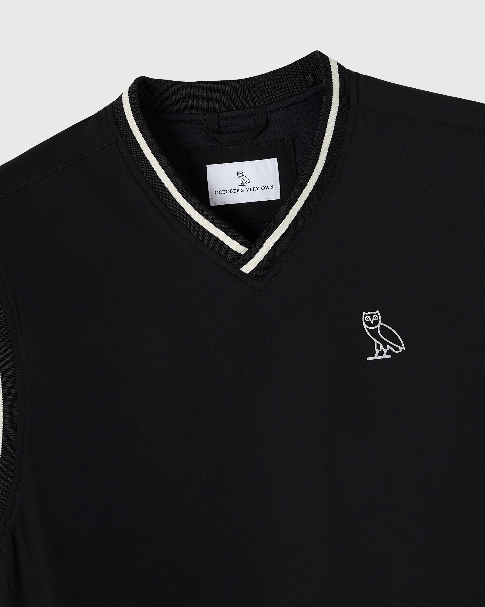 Black pullover vest with white stripe trim at V-neckline and owl logo embroidered on chest.