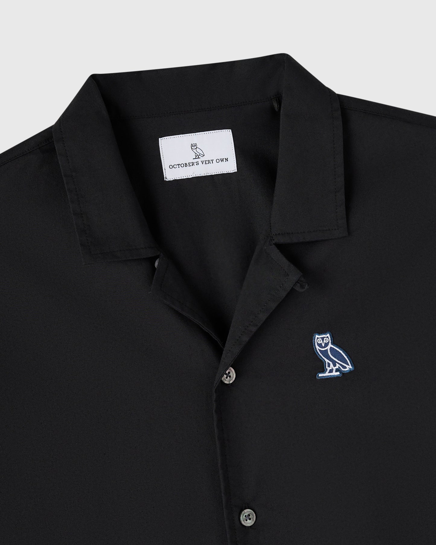 Black camp shirt with notched collar, button front, and embroidered owl logo on chest.