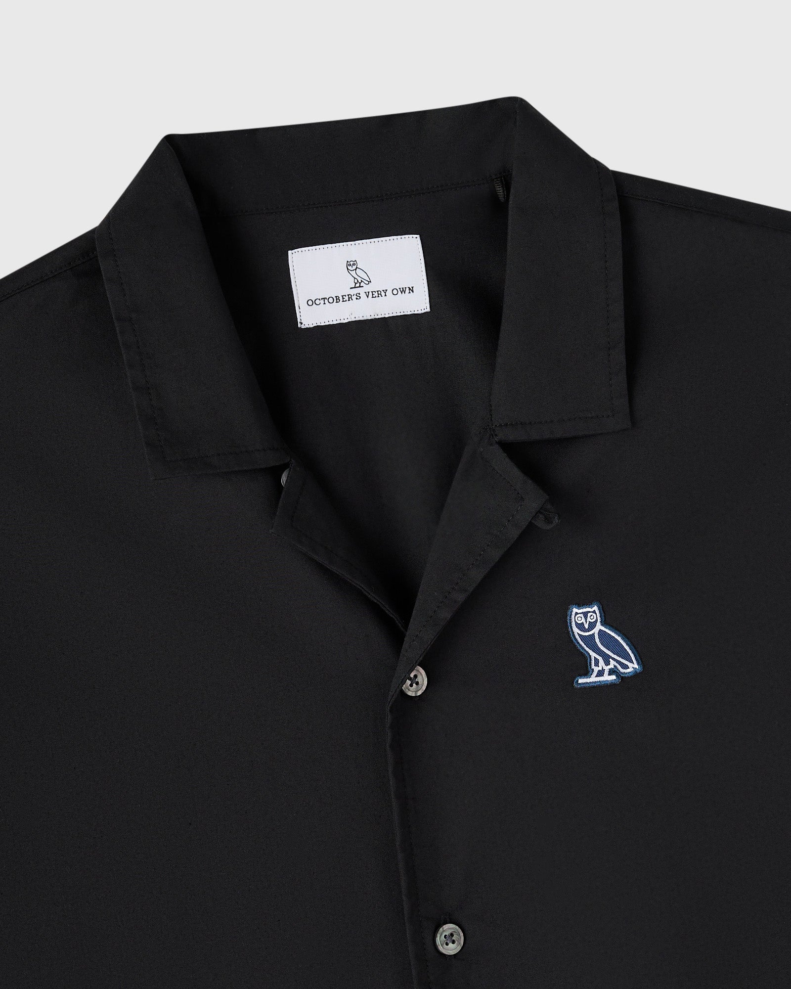 Black camp shirt with notched collar, button front, and embroidered owl logo on chest.