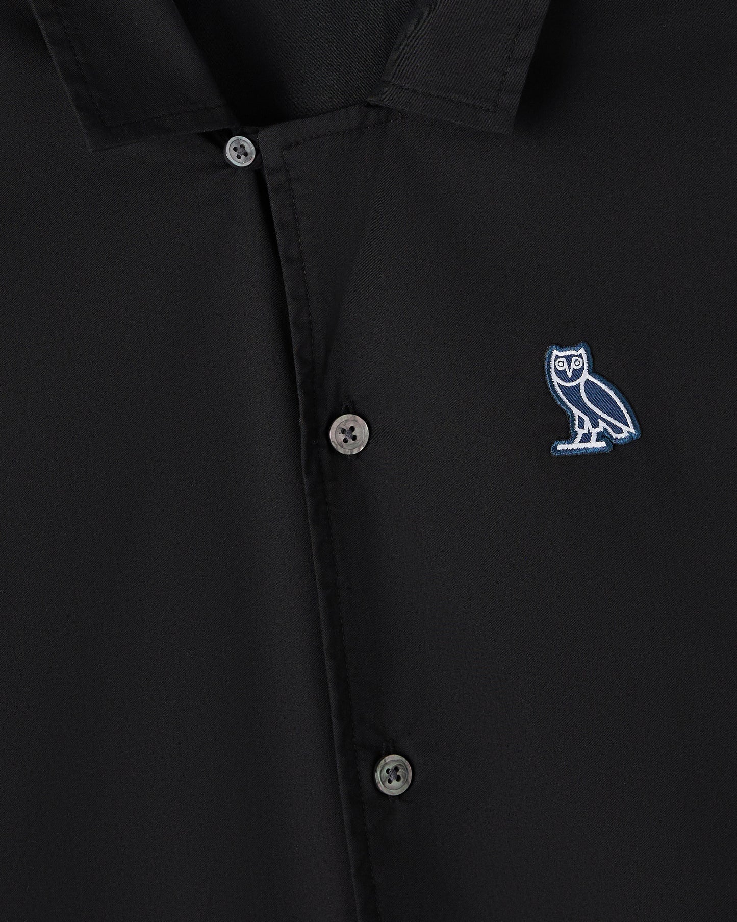 Black camp shirt with button front closure and blue owl embroidered logo on chest.