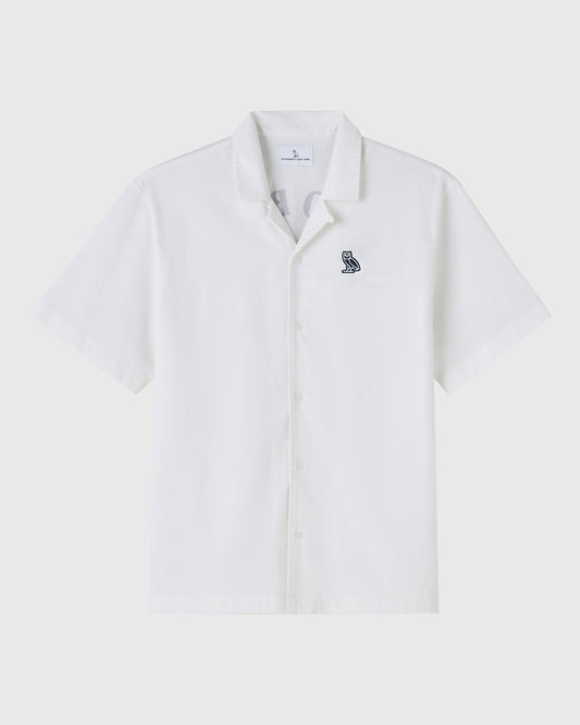 White short-sleeve camp shirt with small embroidered owl logo on chest and button front closure.