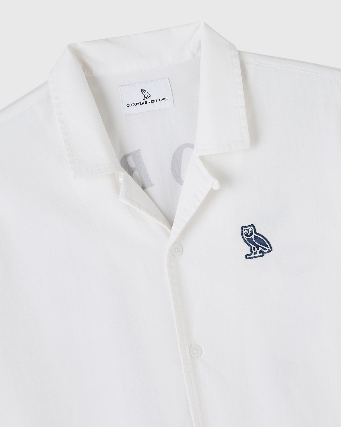 White camp shirt with button front closure and navy owl embroidery on chest.