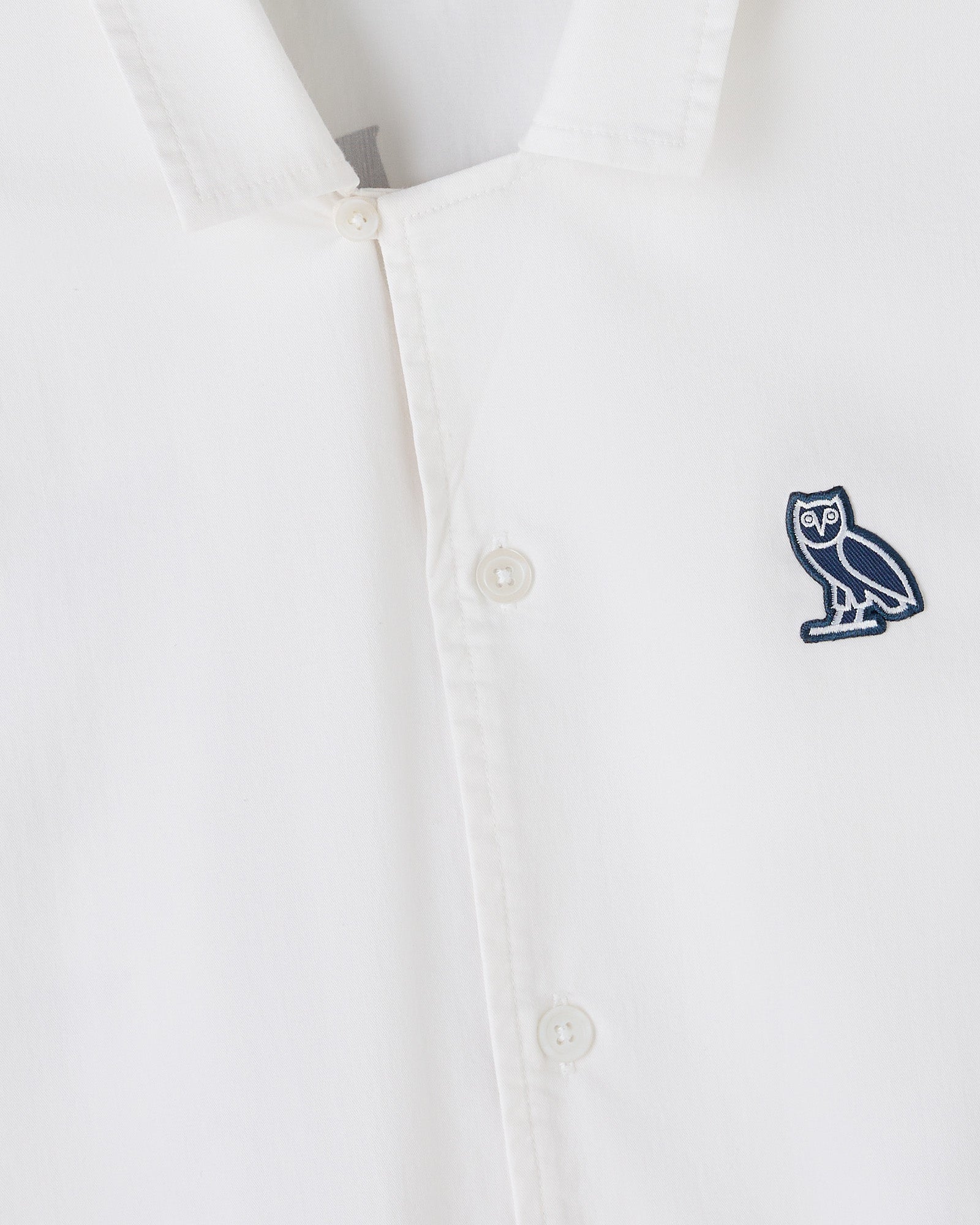 White camp shirt with button front closure and navy owl embroidered patch on chest.
