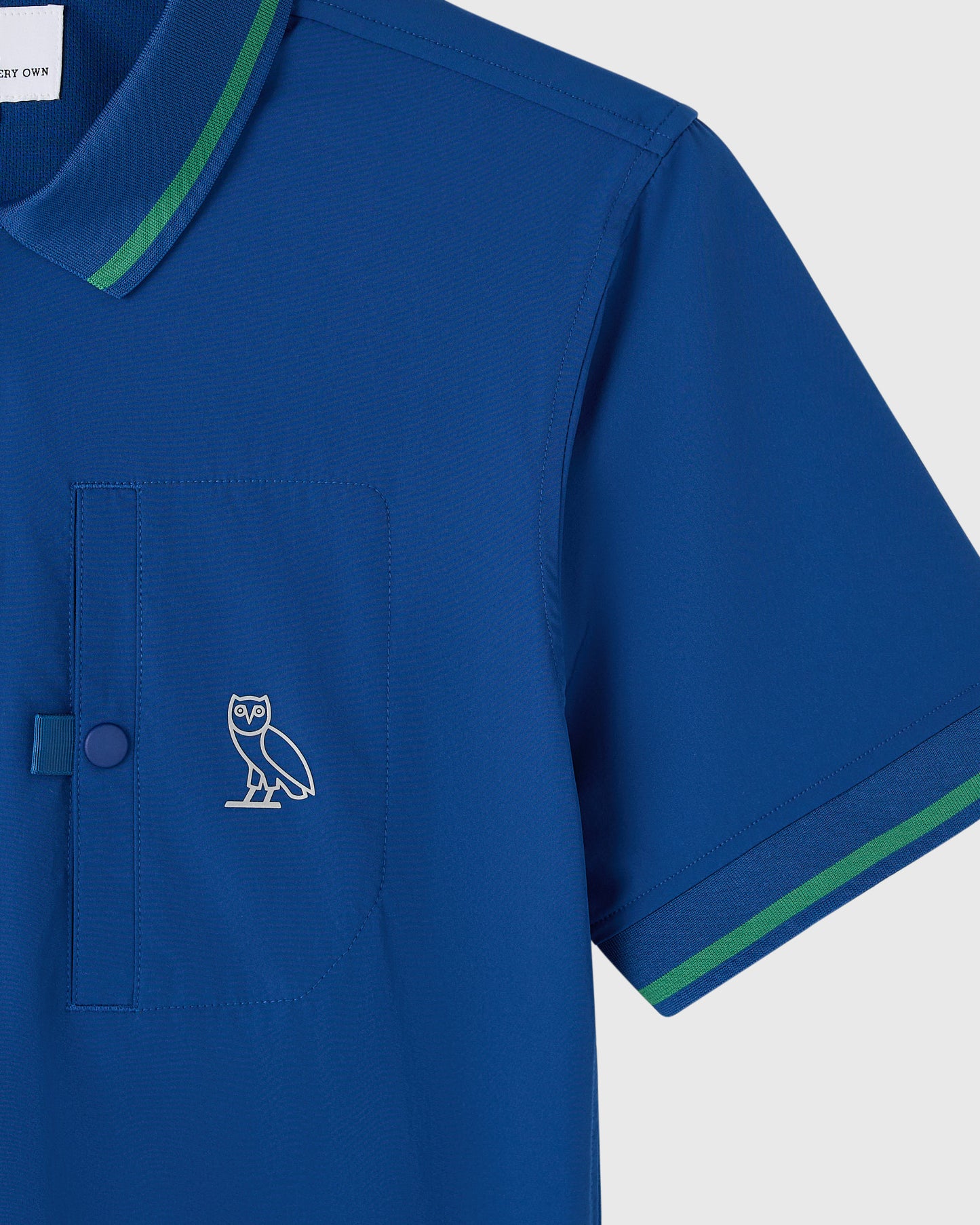 Deep denim polo shirt with owl logo, green trim on collar and sleeves, chest pocket with button.