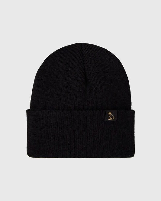 Black ribbed knit beanie with small gold owl emblem on folded cuff.