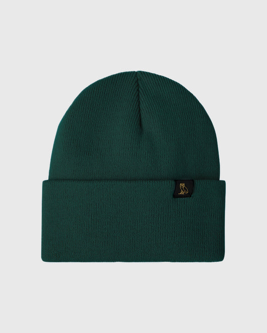 Midnight green ribbed knit beanie with black label patch and folded cuff.