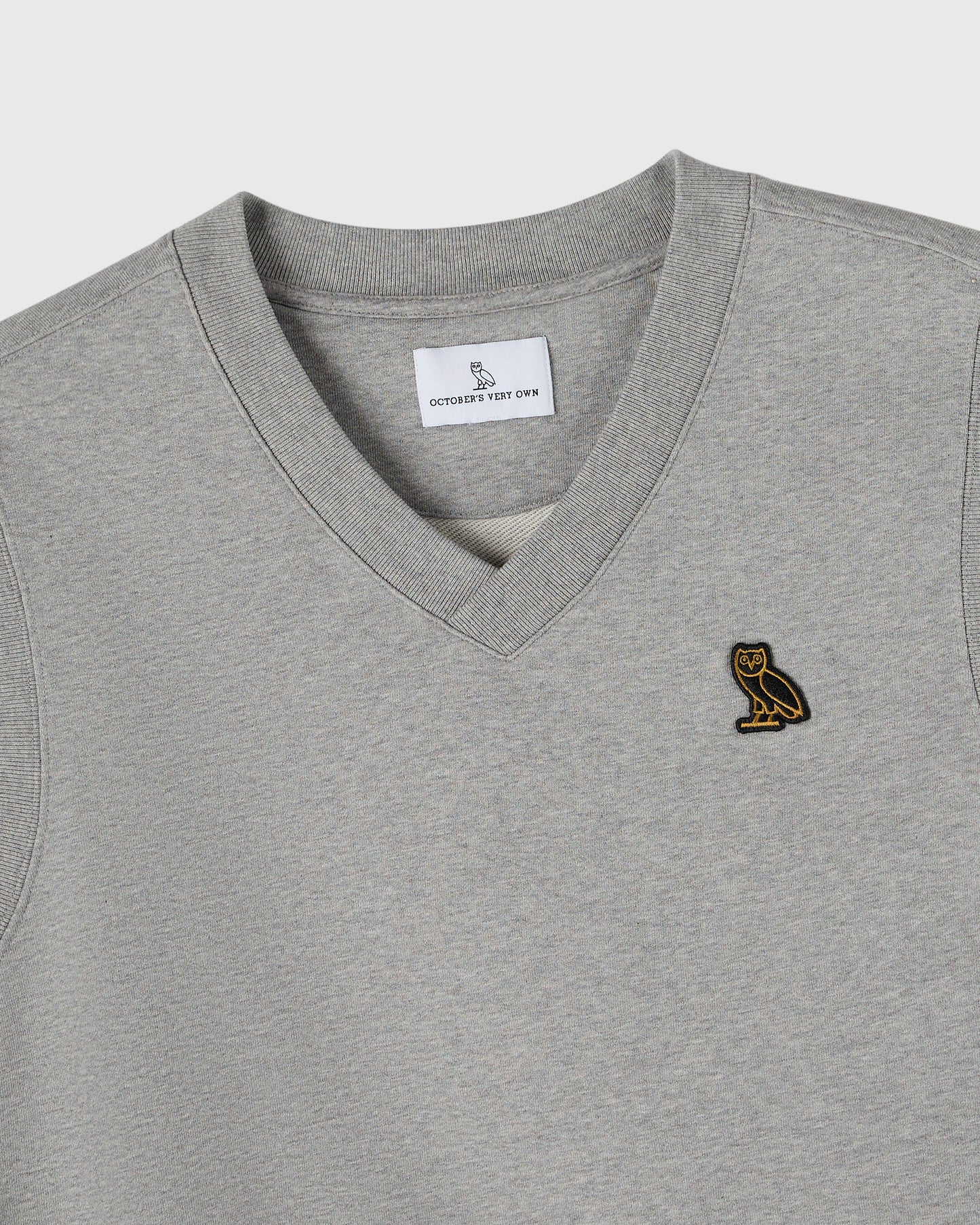 Heather grey v-neck vest with small gold owl embroidered on chest and brand label visible at neckline.