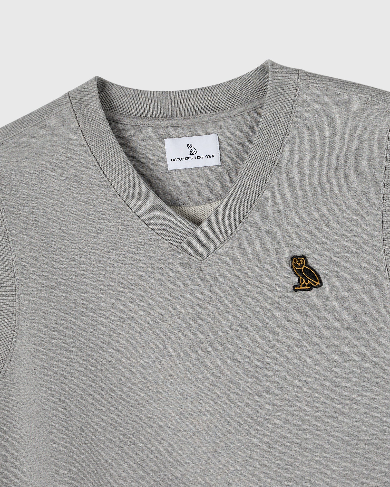 Heather grey v-neck vest with small gold owl embroidered on chest and brand label visible at neckline.