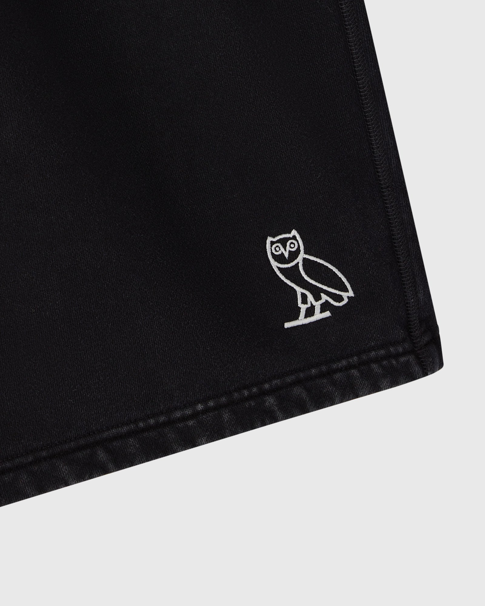 Black sweatshorts with white owl embroidery on the leg and stitched hem detail.