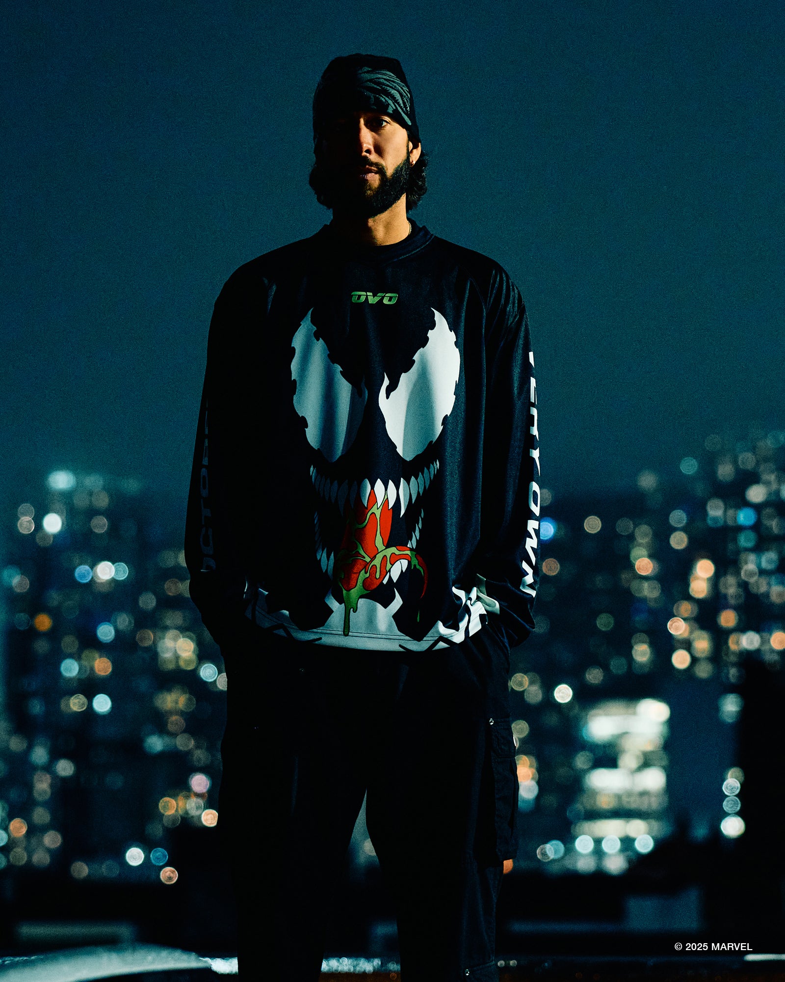 Person wearing a black October's Very Own long-sleeve shirt with a white skull design against a cityscape at night.