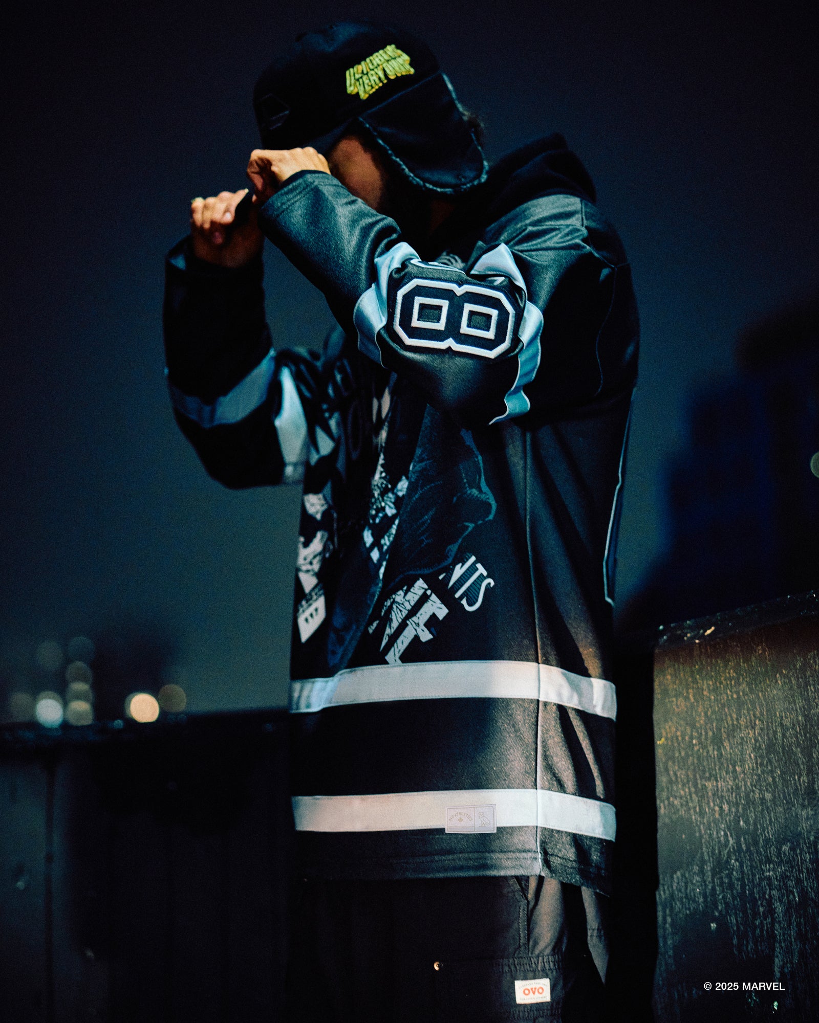 Person wearing a dark October’s Very Own hoodie with reflective stripes and a logo, standing against a dark background.