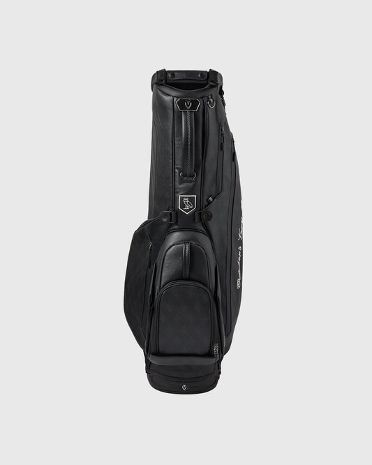 Black Vessel Monogram Lux stand bag with Vessel logo badge, front pockets, and shoulder strap on light background.