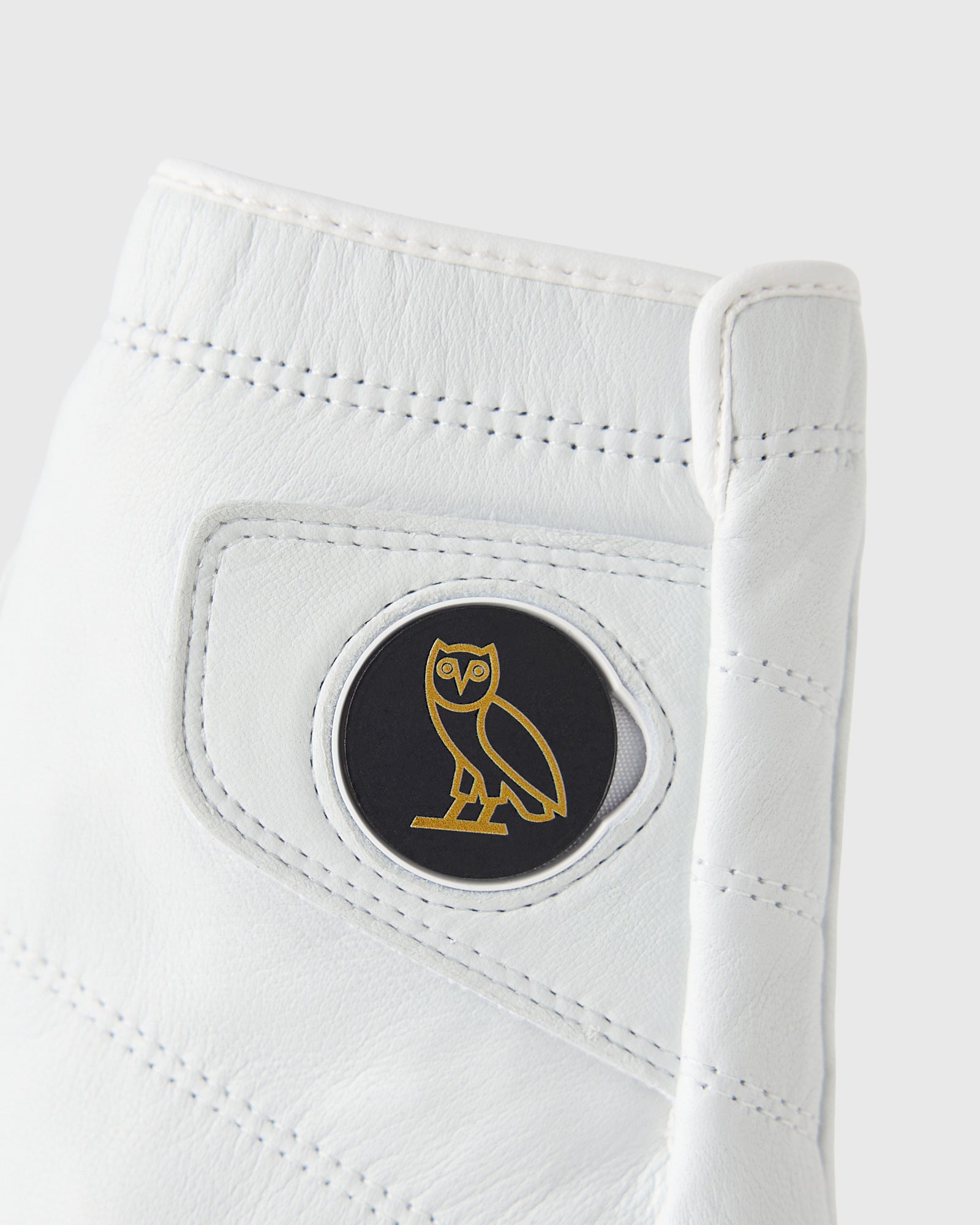 White leather golf glove with perforated details and circular black and gold owl logo on palm.