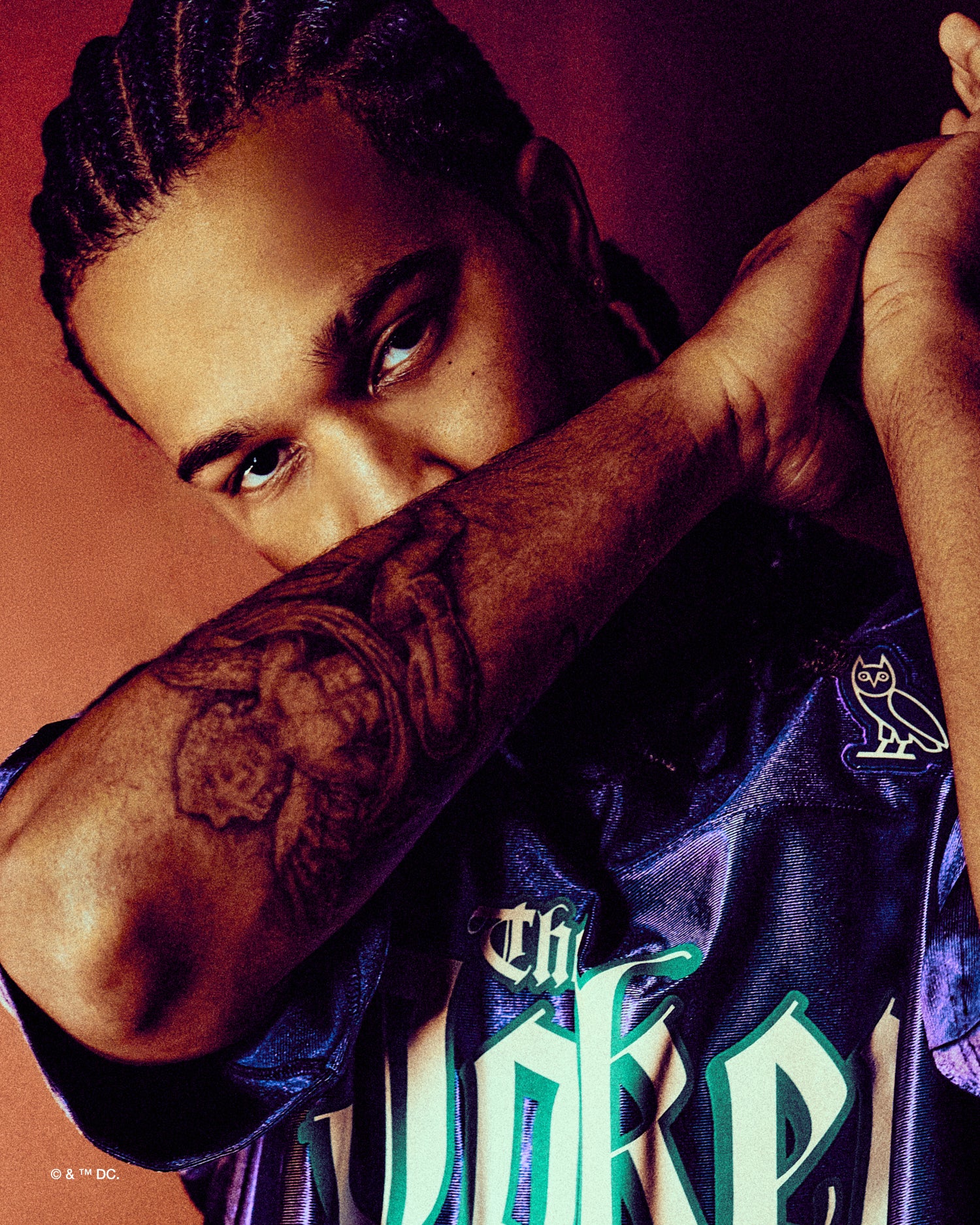 Person wearing a purple October's very Own x Batman x The Joker sports jersey with visible tattoos on an orange background