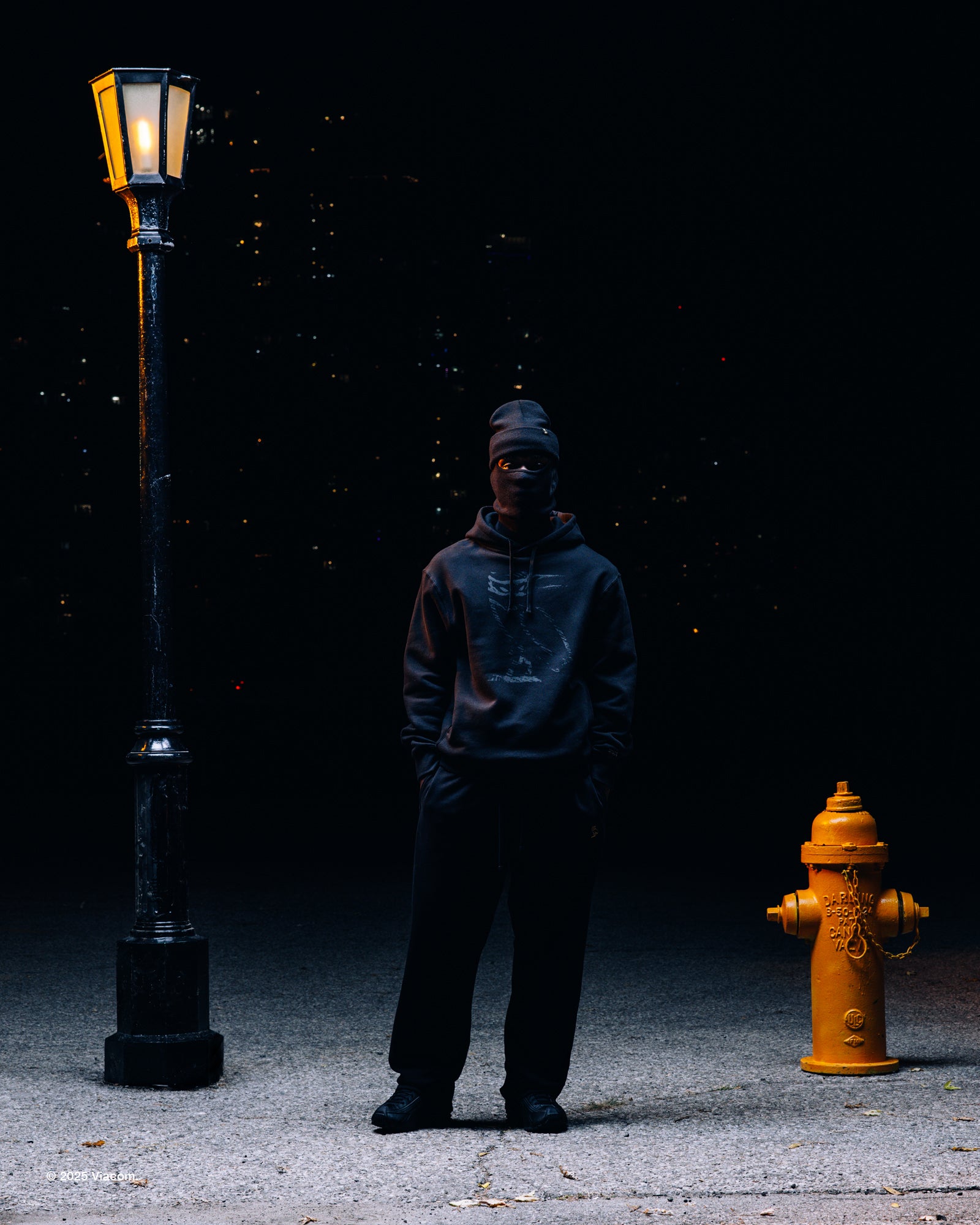 Person wearing a x Very Own x TMNT hoodie and mask standing between a street lamp and fire hydrant on a dark street.