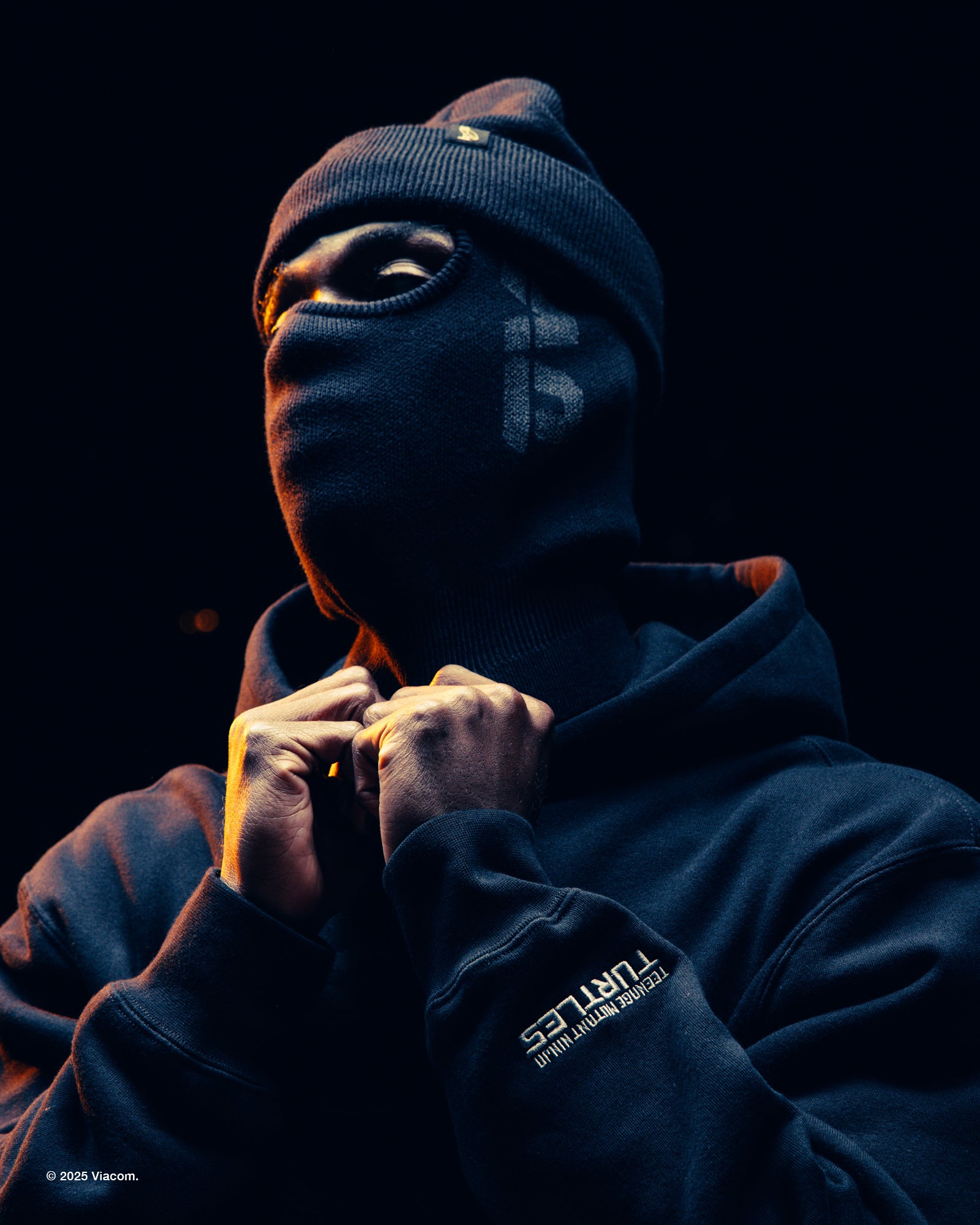 Person wearing a dark October's Very Own x TMNT hoodie and balaclava with a visible brand logo on a black background