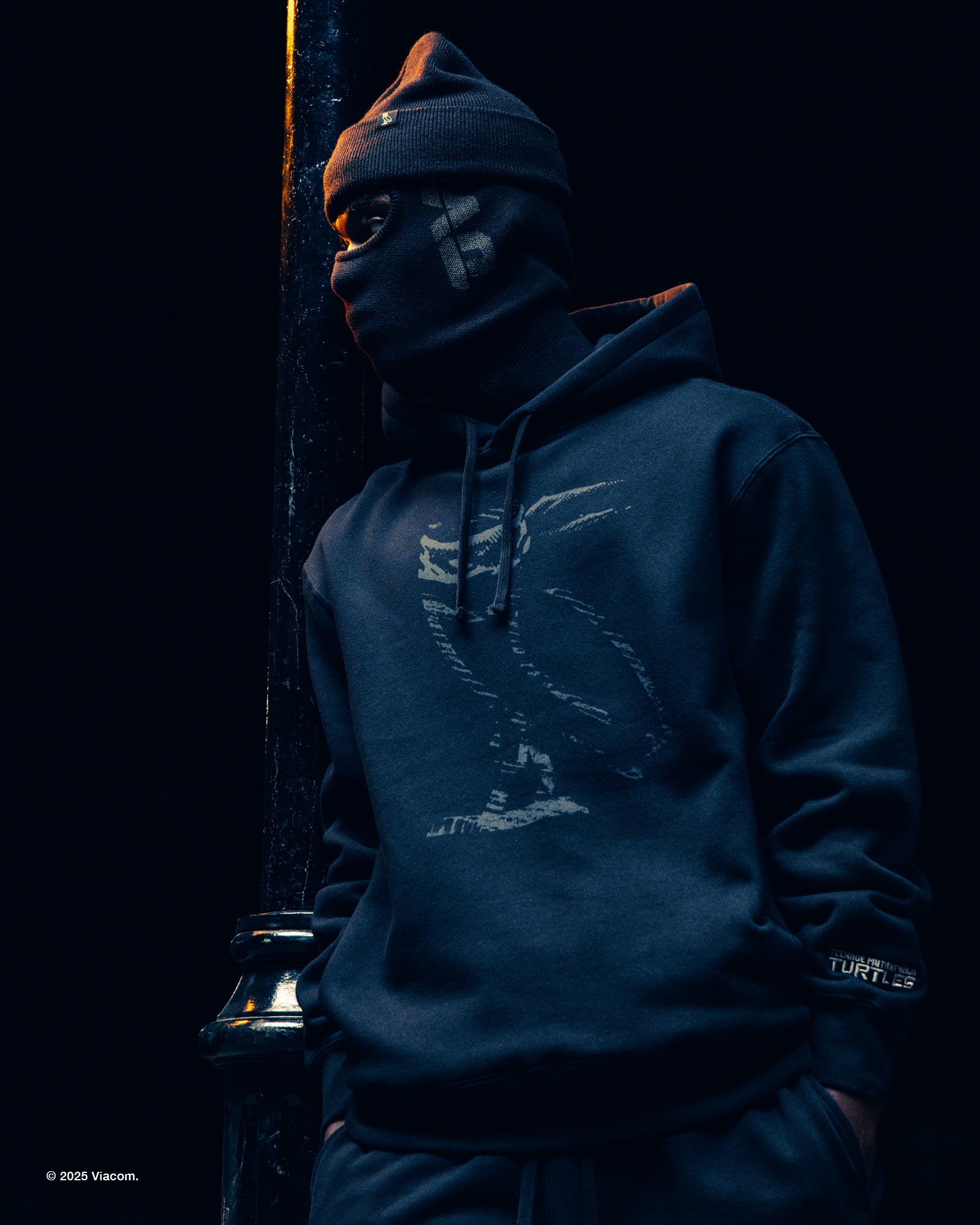 Person wearing a dark blue October's Very Own x TMNT hoodie with a logo, standing against a dark background.