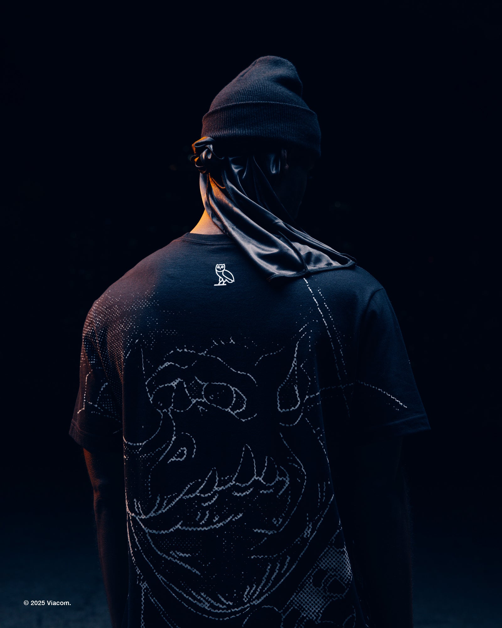 Person wearing a black October's Very Own x TMNT t-shirt with a logo and design, standing against a dark background.
