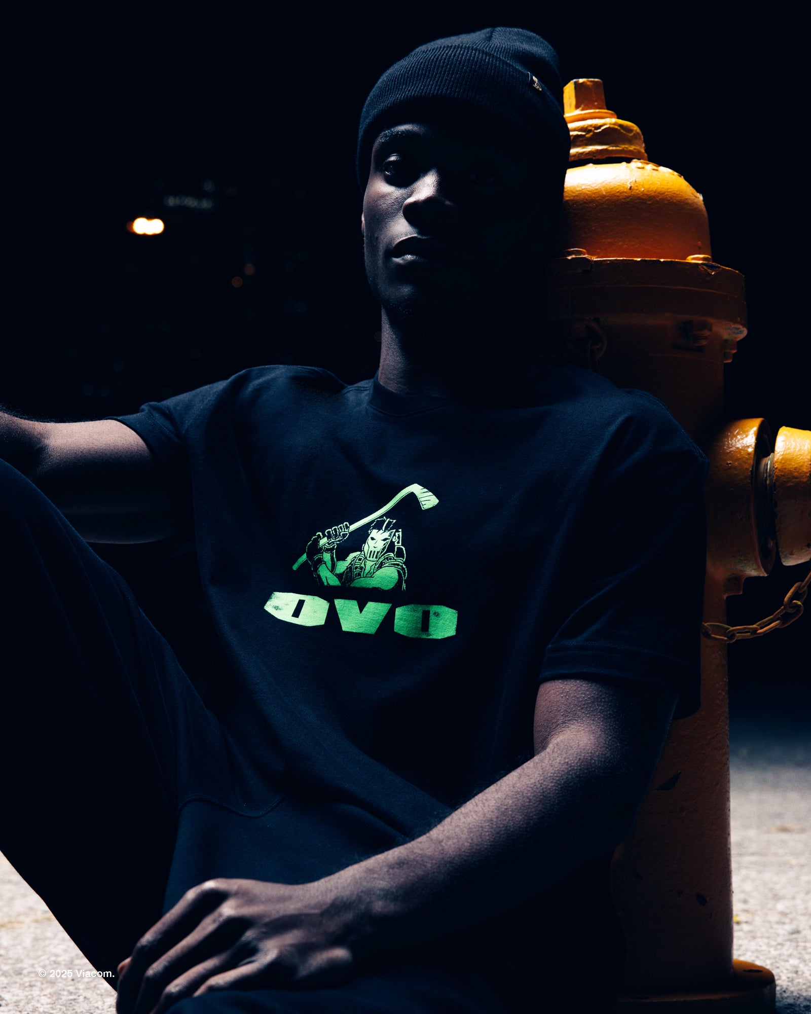 Person wearing a black October's Very Own x TMNT t-shirt with a green logo sitting against a dark background