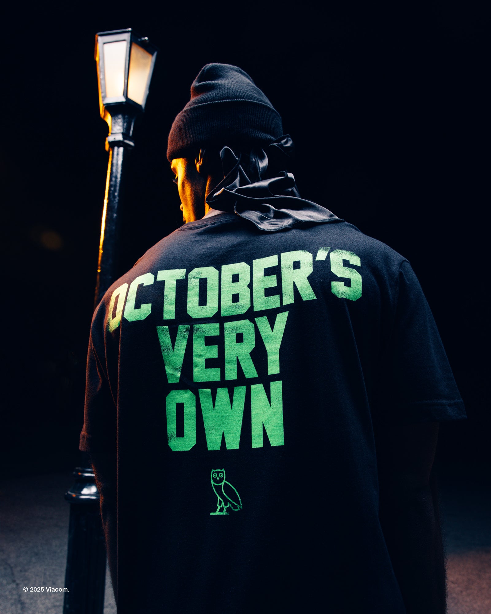 Person wearing a black hoodie with 'October's Very Own' text in green, standing in front of a street lamp at night.