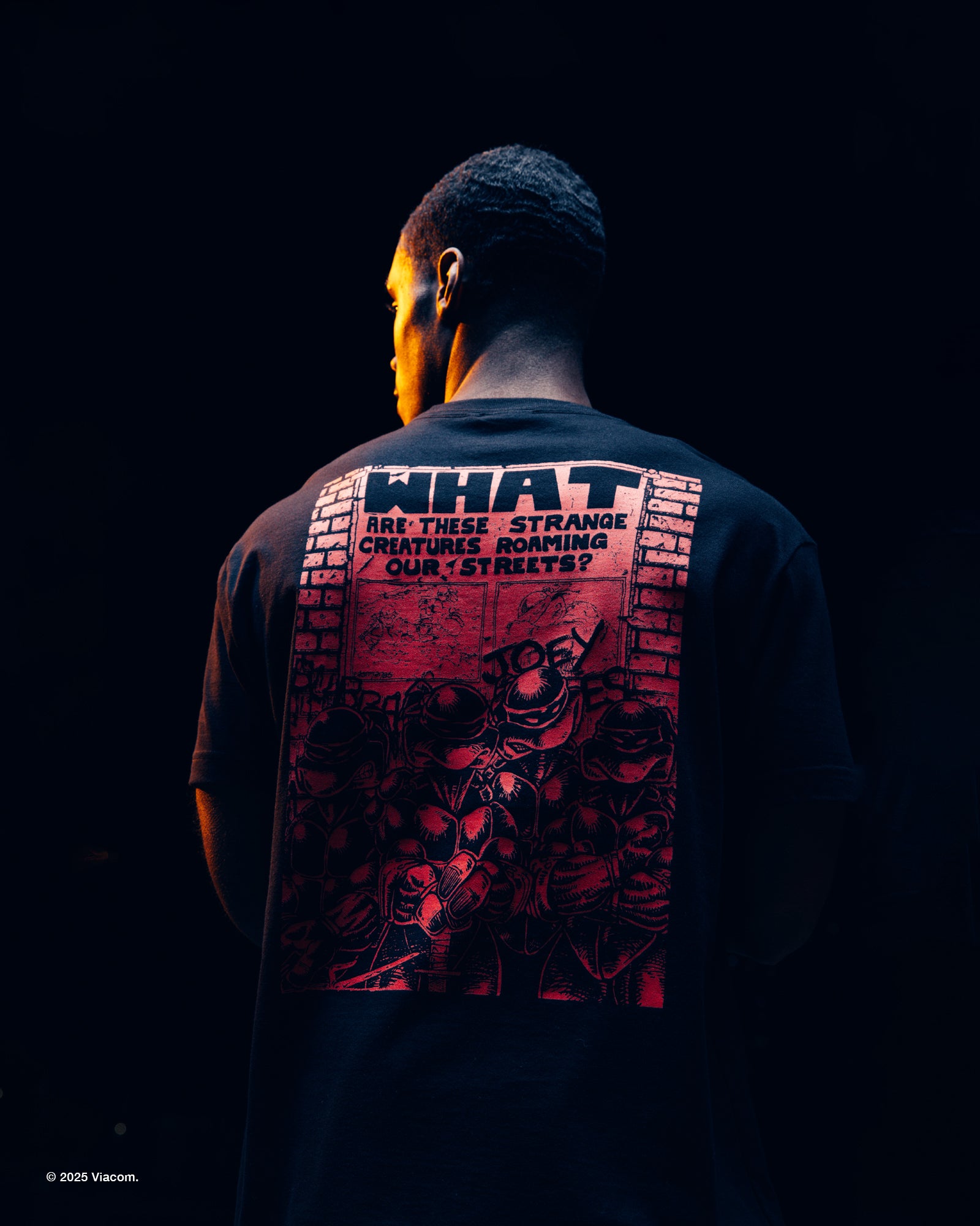 Person wearing a black October's Very Own x TMNT t-shirt with a graphic design on the back, set against a dark background.