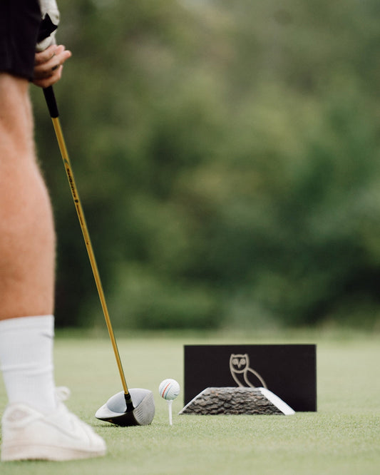 Golfer holding club above white ball on tee with OVO logo marker on grass course.