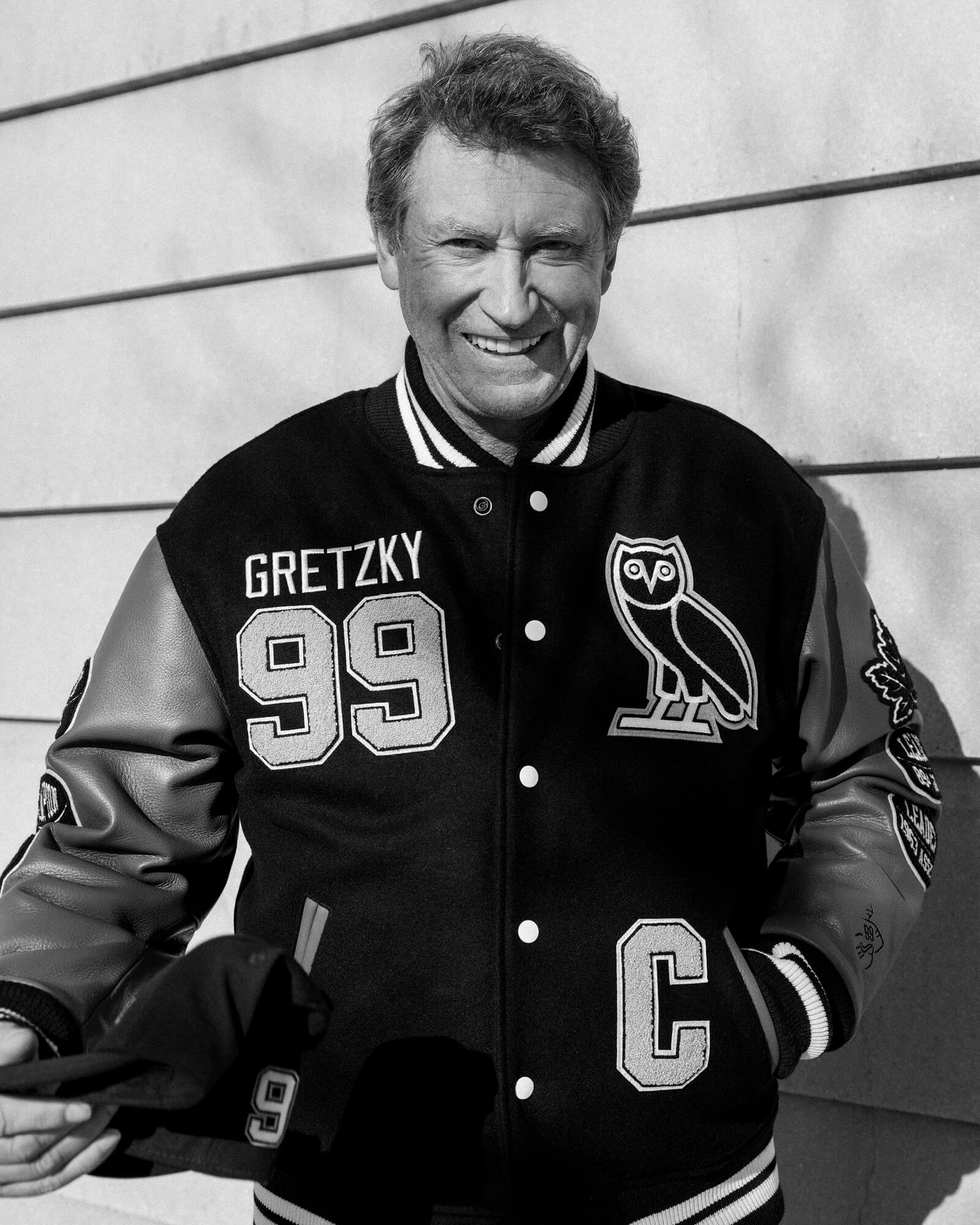 Wayne Grezky wearing a October's Very Own Varsity jacket with 'Gretzky 99' and an owl logo, standing against a wall.