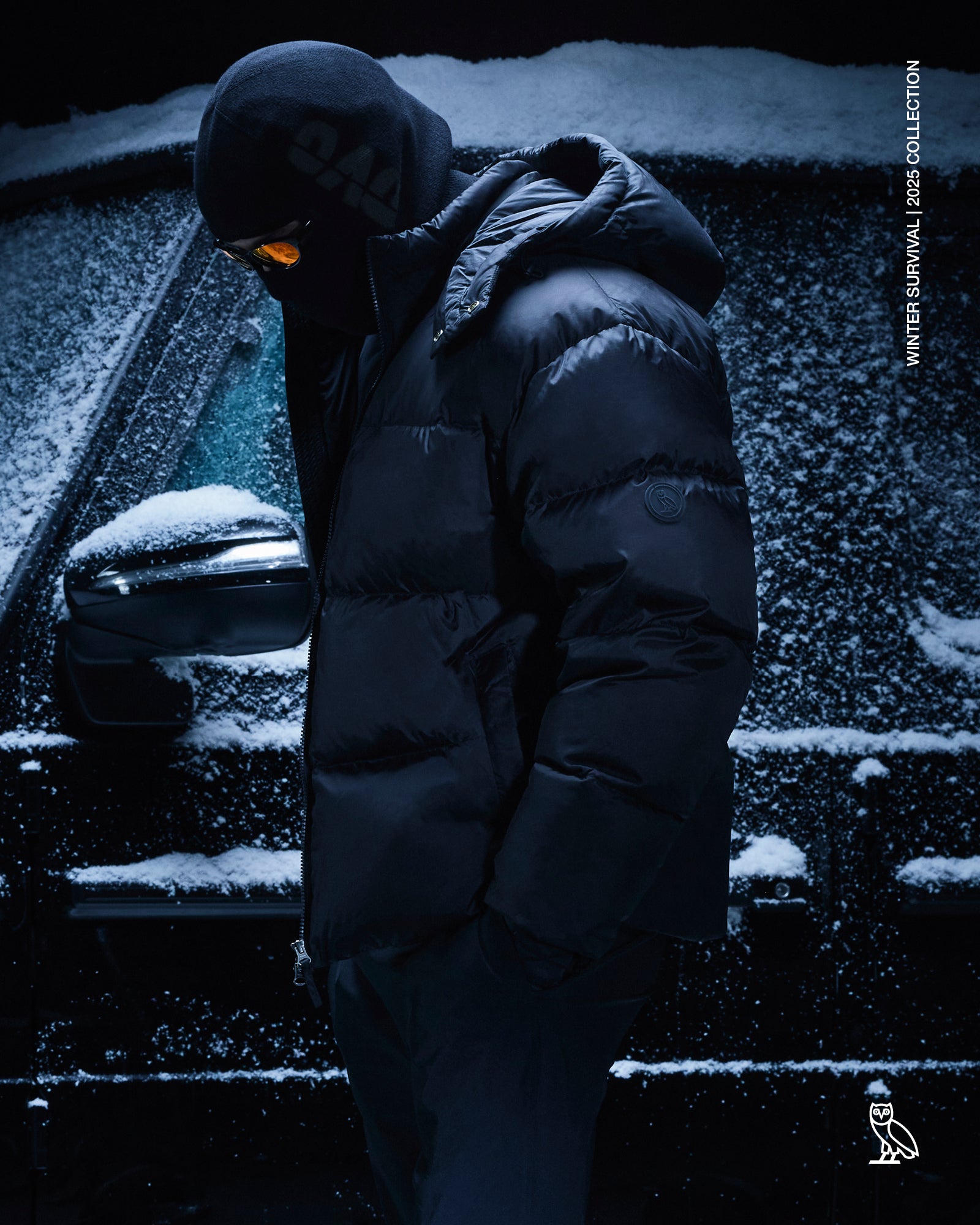Person wearing a October's Very Own  black puffer jacket standing next to a snow-covered car at night.