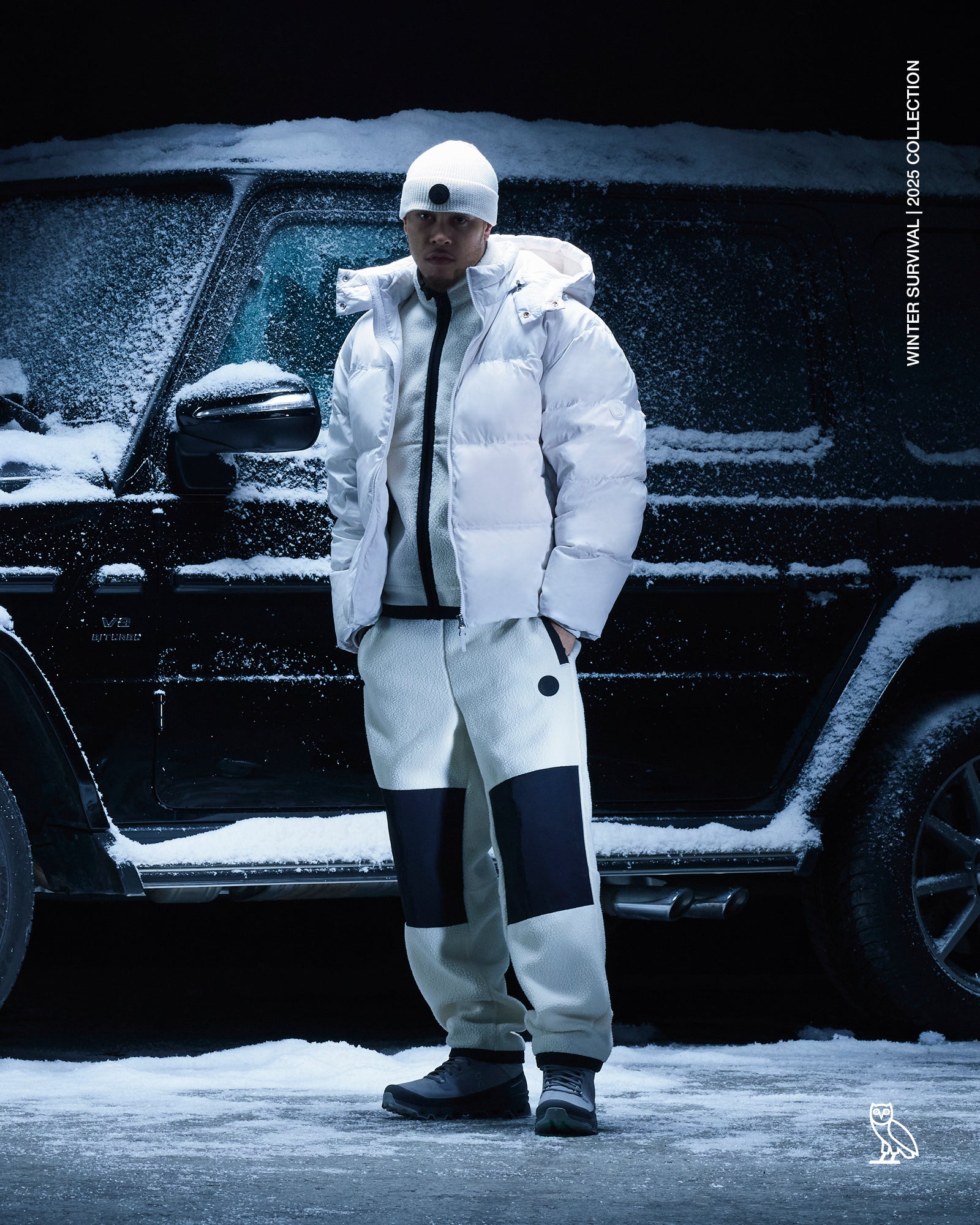 Person wearing a October's Very Own white winter coat and pants standing in front of a snow-covered vehicle.
