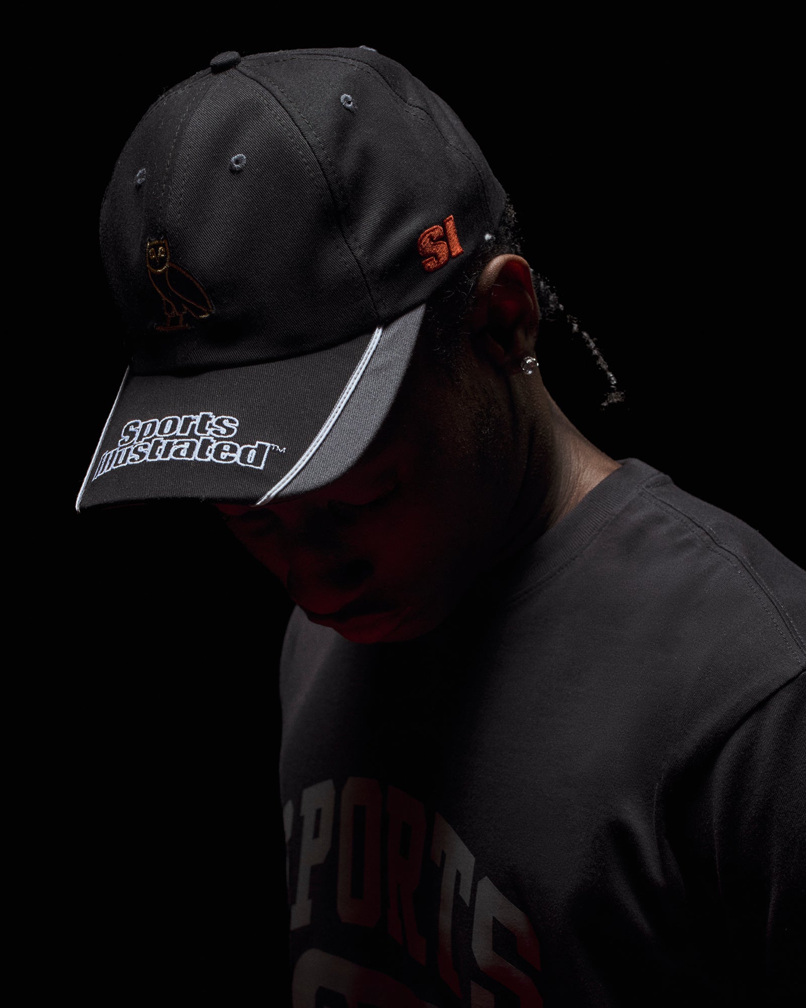 Person wearing a black October's Very Own x Sports Illustrated cap with visible branding against a dark background